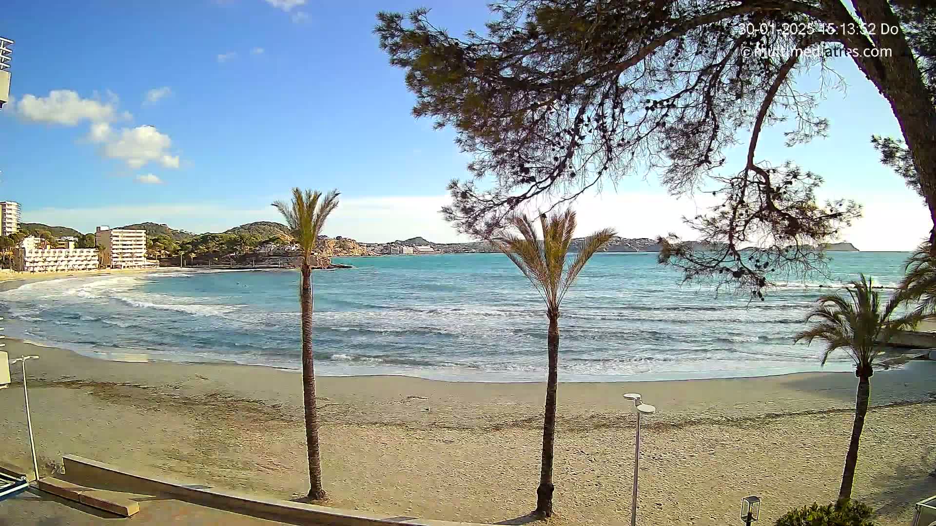 A sandy beach with gentle waves laps the shore under a mostly sunny sky, palm trees and hotel buildings are visible in the background.