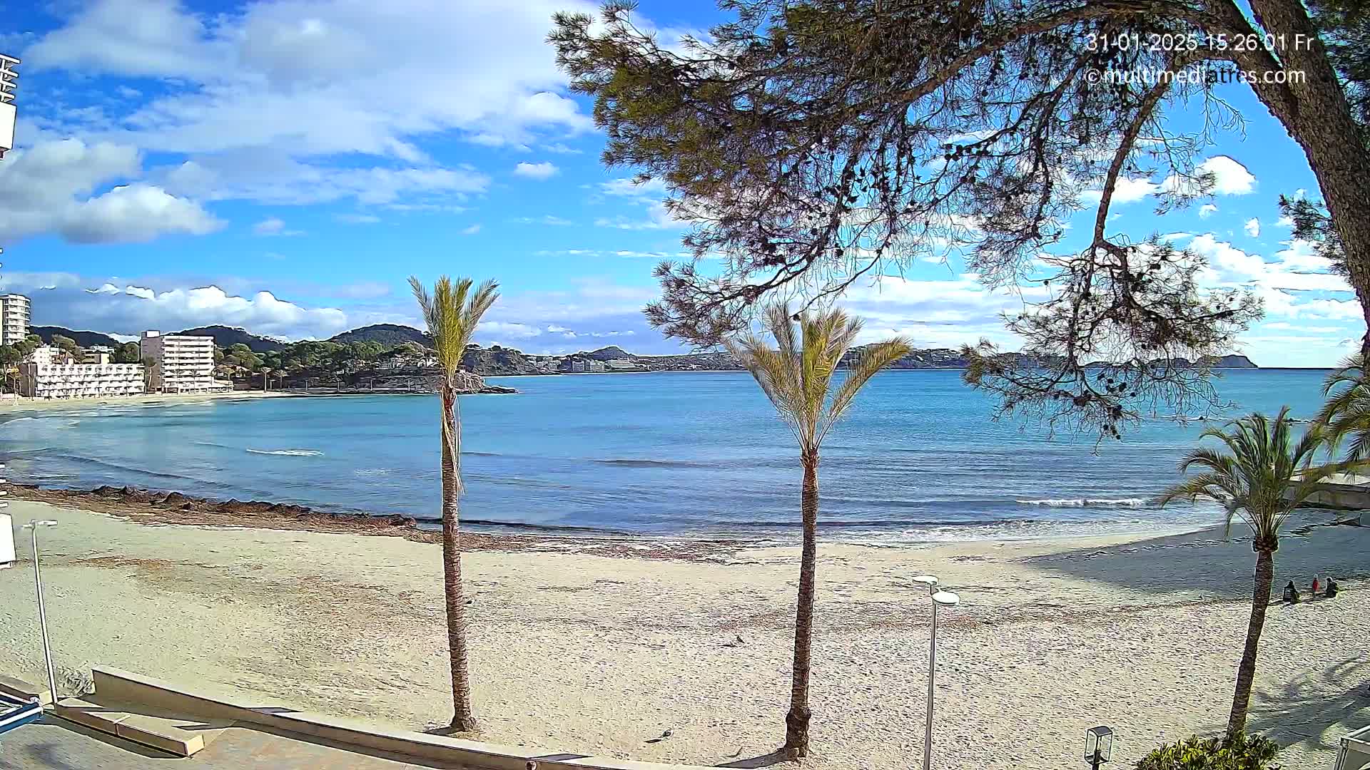 A partly cloudy sunny day shows a sandy beach with palm trees, ocean waves, and hotel buildings in the distance.