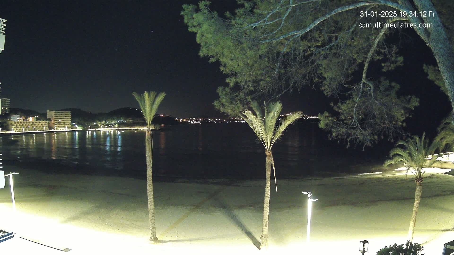 A nighttime view of a sandy beach, calm water, and a town across the bay illuminated under a clear, starry sky.