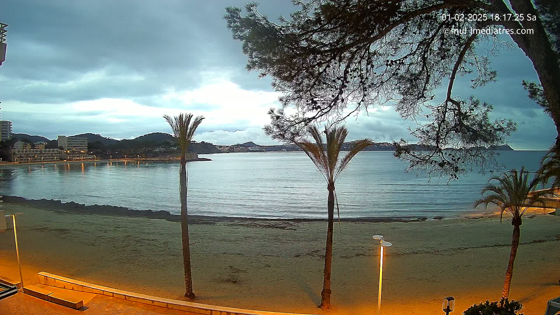 A sandy beach at dusk or dawn, under a cloudy sky, with calm water and palm trees in the foreground, overlooking buildings and hills in the distance.