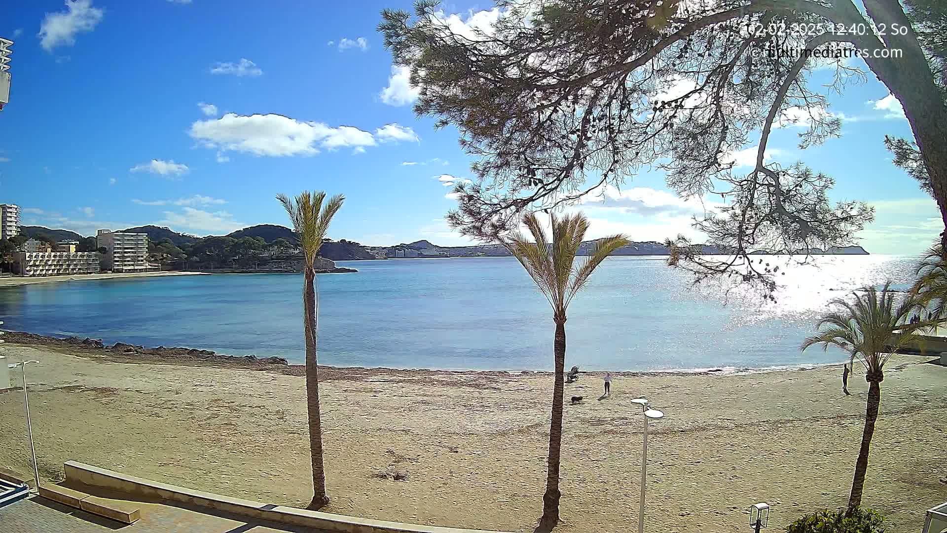 A sandy beach with palm trees and buildings in the background stretches along a calm, turquoise bay under a mostly sunny sky with some clouds.