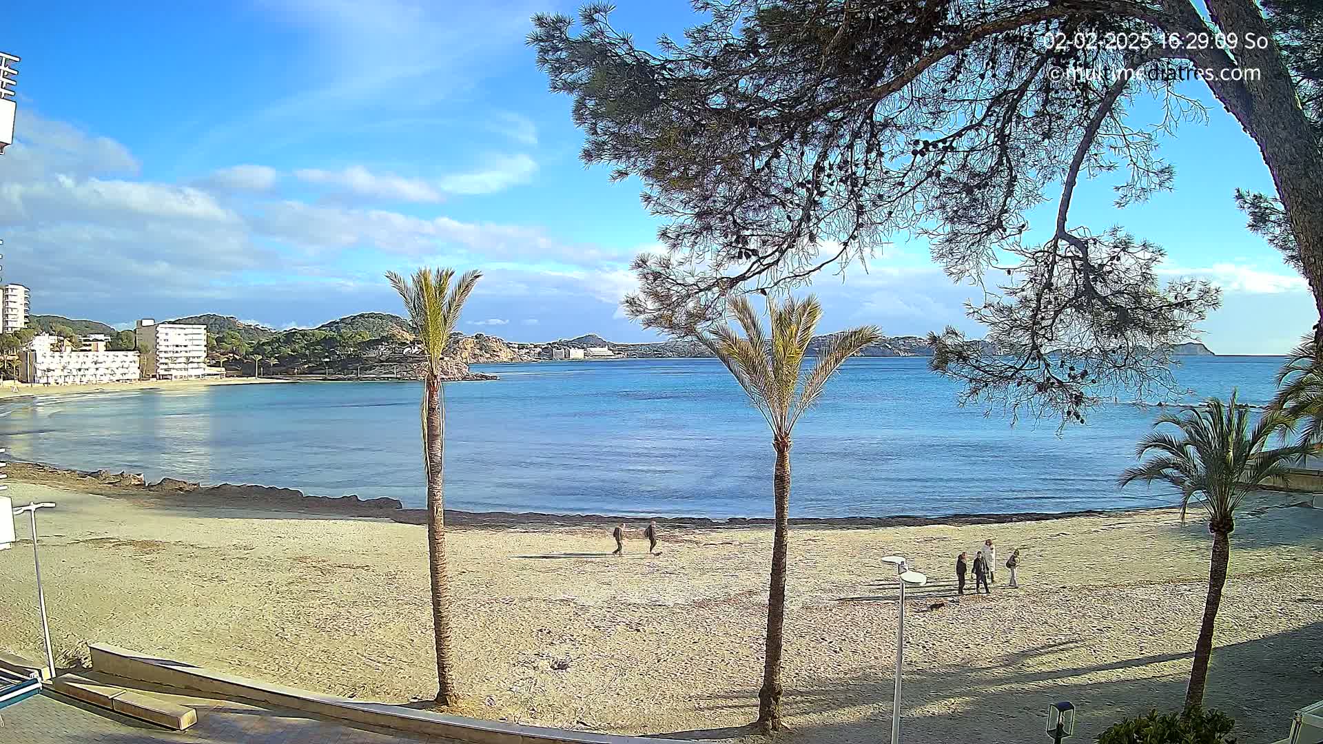 A mostly sunny day shows a sandy beach with several people walking, palm trees, and calm blue water with buildings visible in the distance.