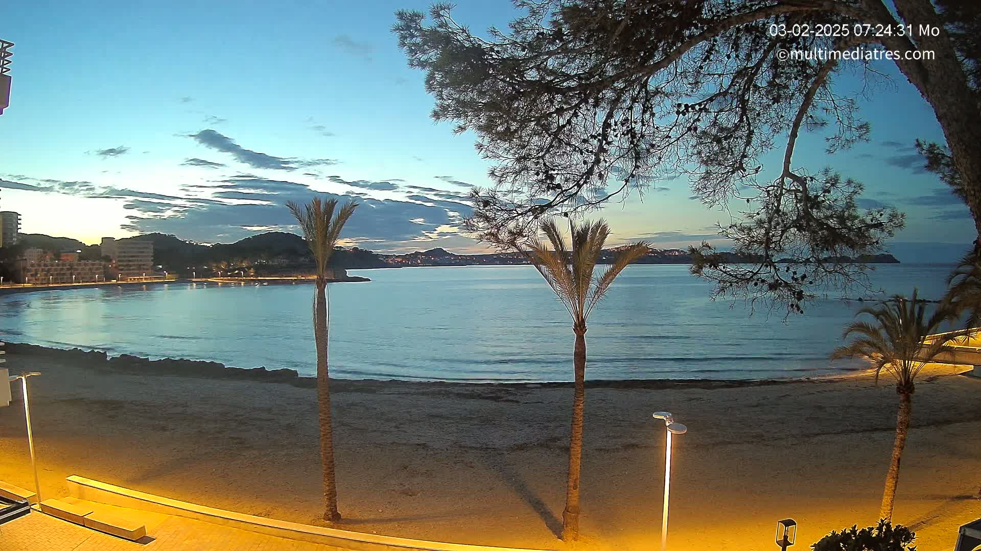 A sandy beach at twilight is bordered by calm ocean water, buildings, and palm trees under a mostly clear sky.
