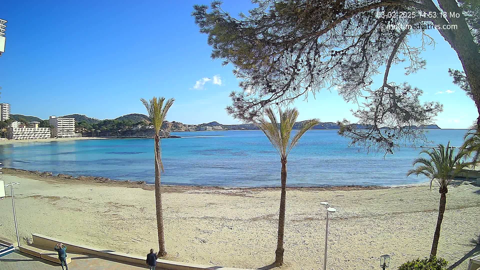 A sandy beach with calm, turquoise water, palm trees, and hotel buildings in the background under a clear blue sky.