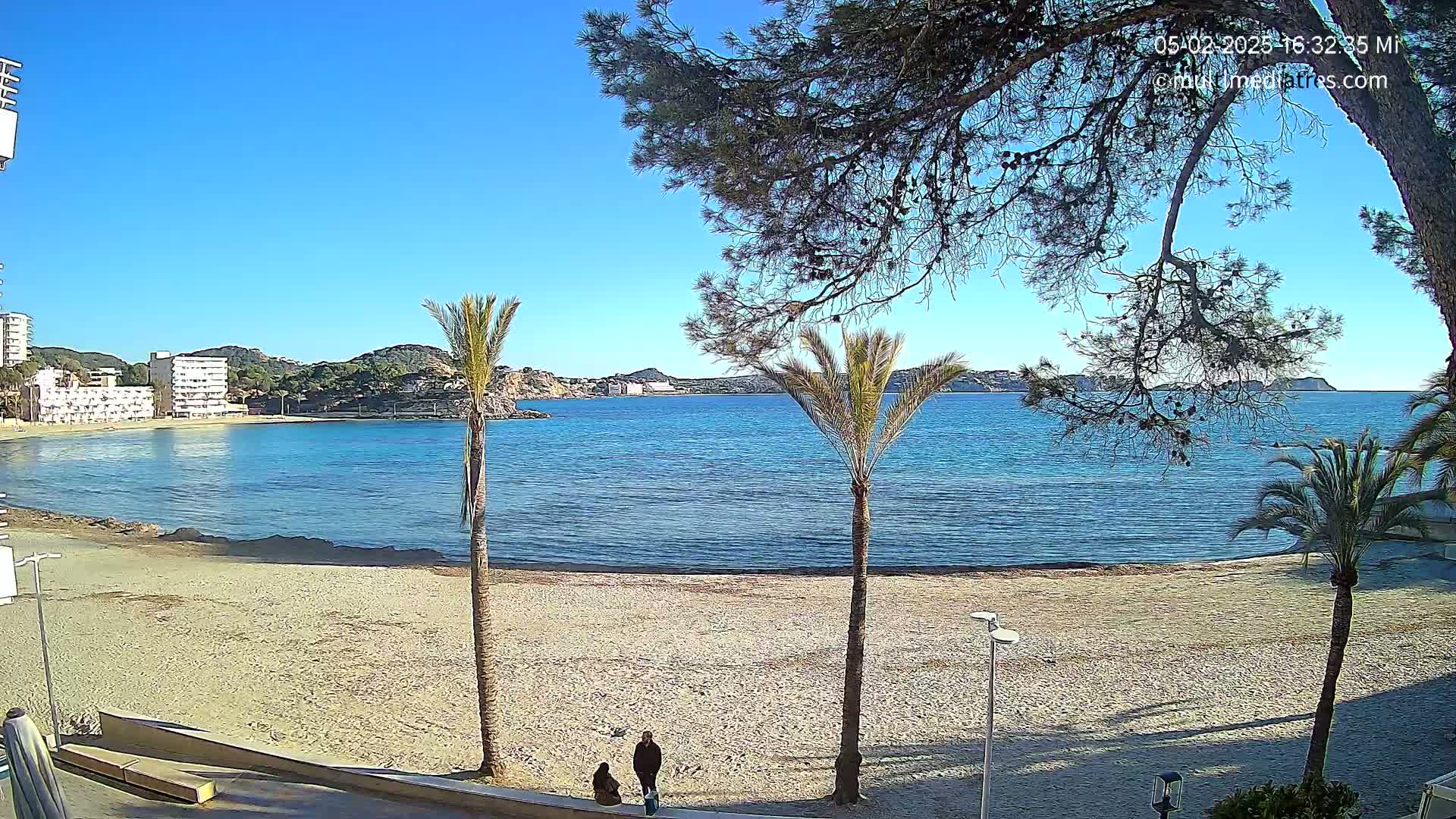 A sandy beach with calm blue water, palm trees, and two people walking is visible under a clear blue sky.