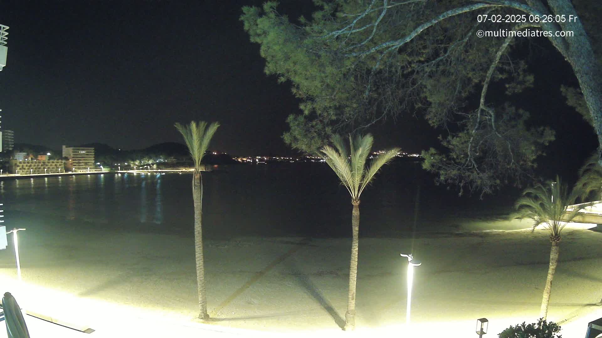 A nighttime view of a sandy beach with palm trees, illuminated by lights, overlooking a calm body of water and a distant, dimly lit town.