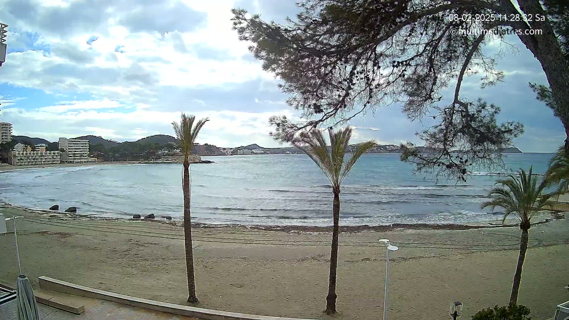 A partly cloudy day shows a sandy beach with gentle waves lapping the shore, palm trees, and buildings in the distance.