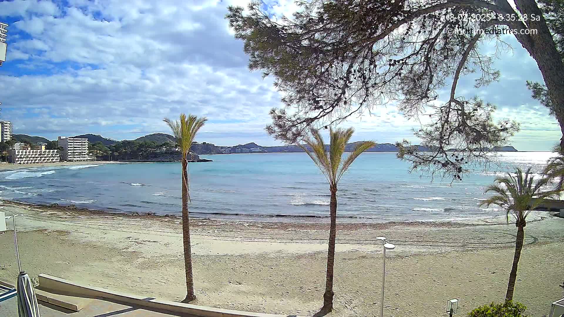 A sandy beach with palm trees and calm, turquoise water under a partly cloudy sky is visible, with buildings and hills in the background.