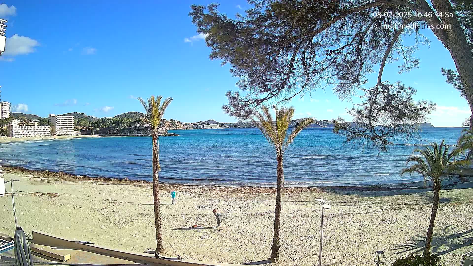 A sandy beach with calm, turquoise water, a few people, and palm trees under a clear blue sky.