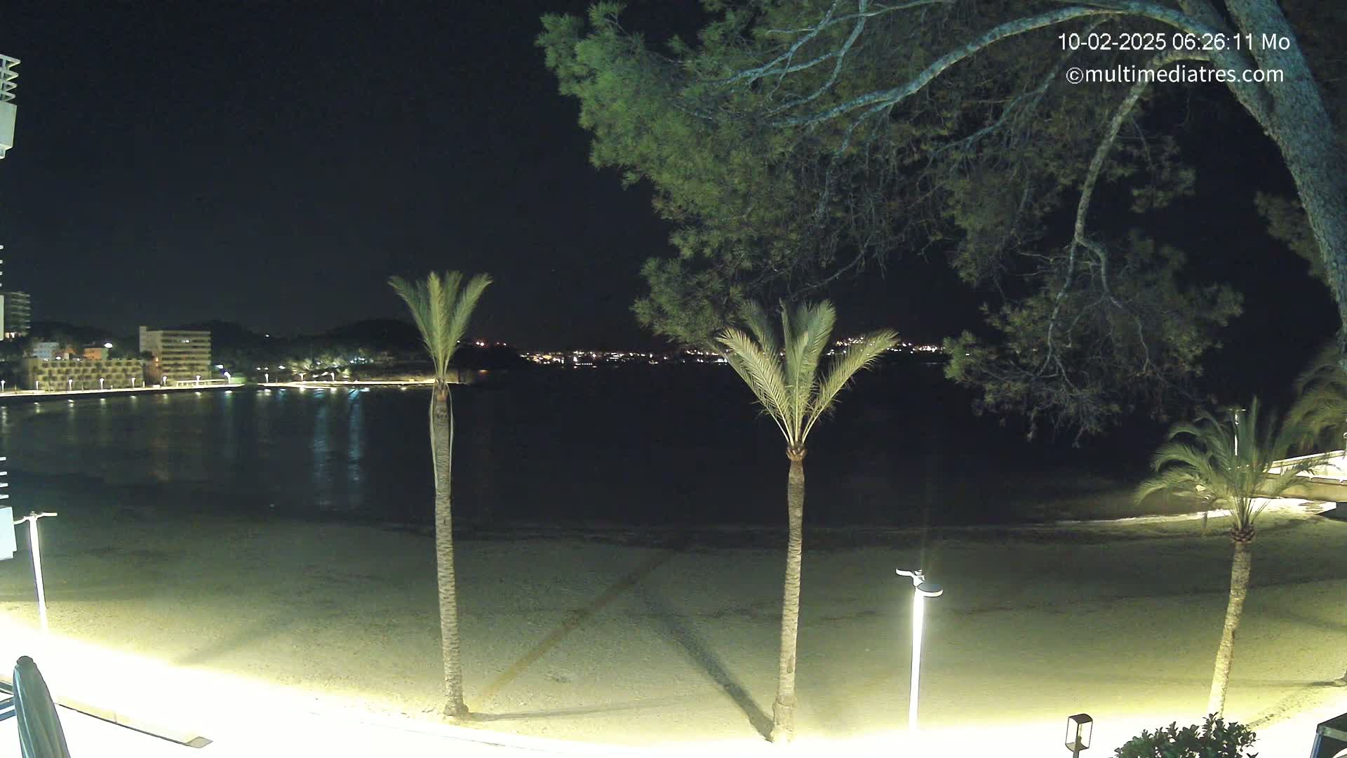 A nighttime view of a sandy beach, calm water, and a town across the bay, illuminated by lights under a clear, dark sky.