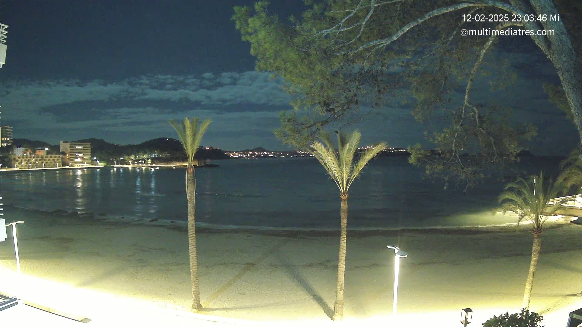 A nighttime view of a sandy beach, calm water, and a city skyline across the bay under a mostly clear sky.