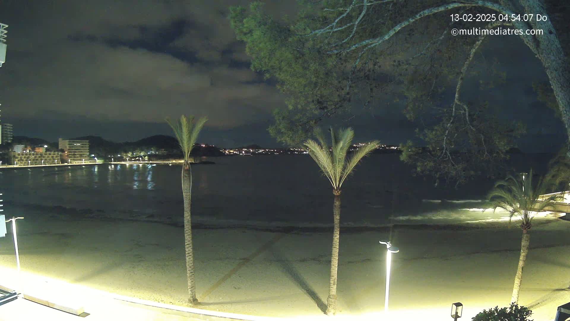 A nighttime beach scene shows a calm sea with small waves lapping the shore under a cloudy sky, illuminated by lights along the beach and buildings across the water.