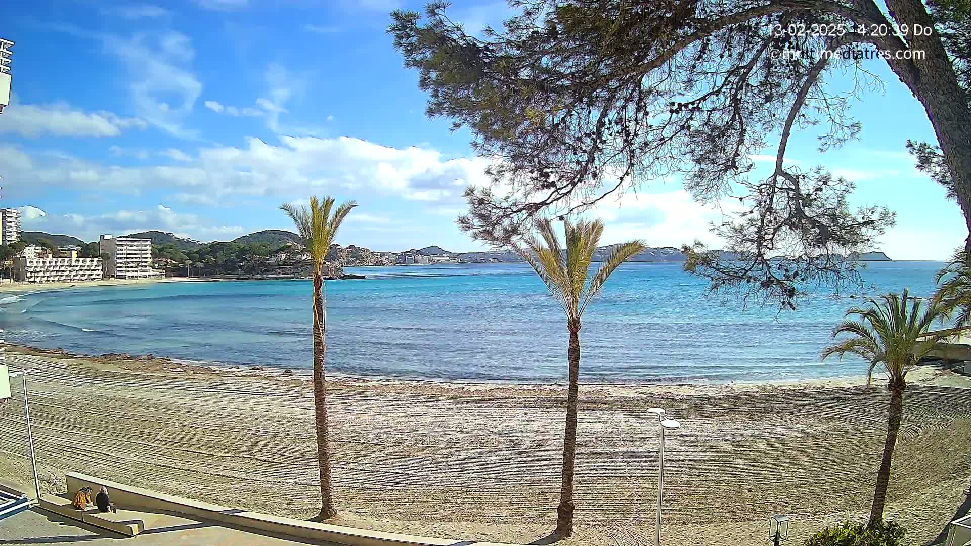 A sandy beach with calm, turquoise water, palm trees, and buildings in the background under a mostly sunny sky with some clouds.