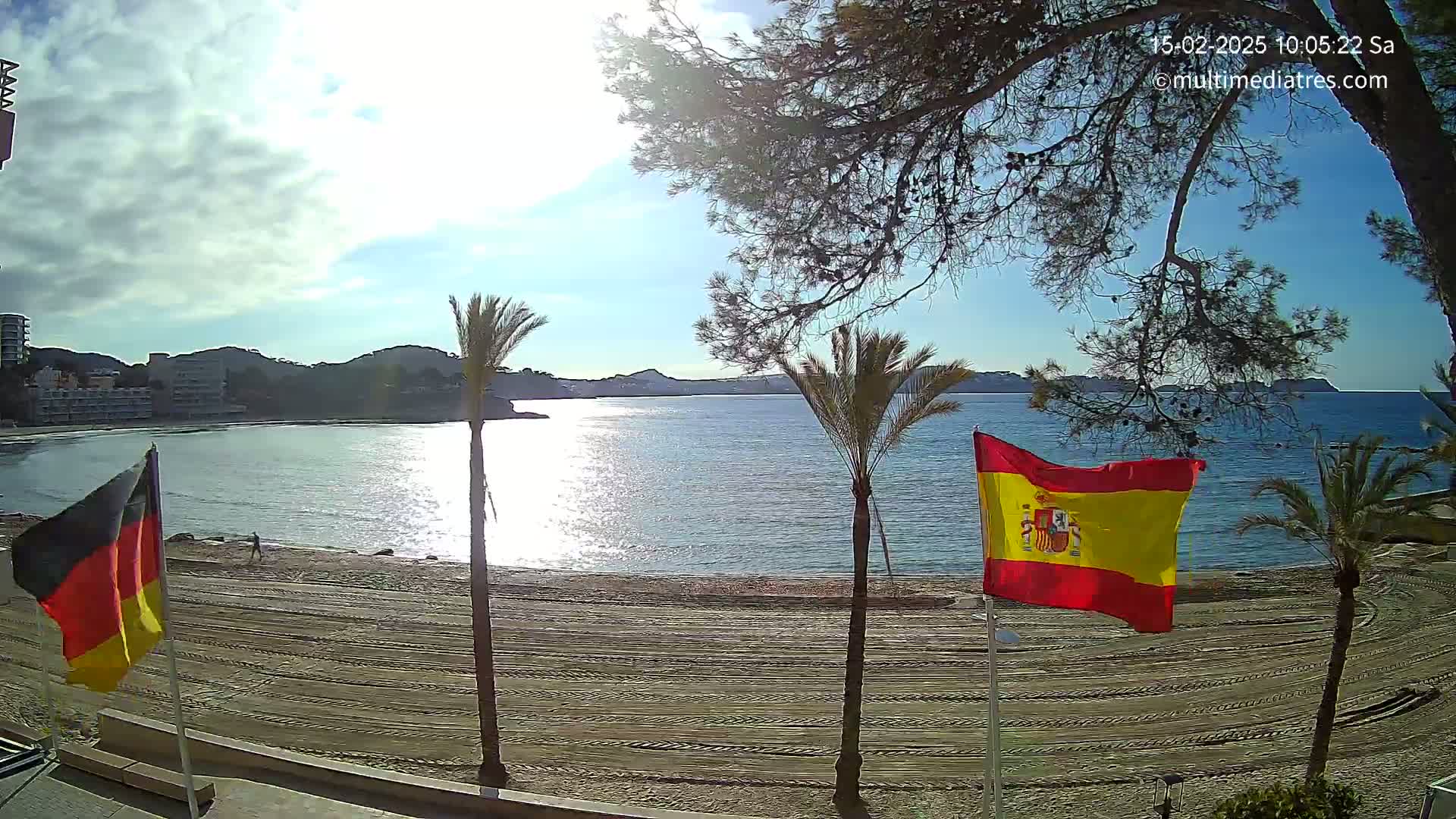 A sunny day on a sandy beach shows a calm bay with palm trees, and a Spanish flag and German flag waving gently.