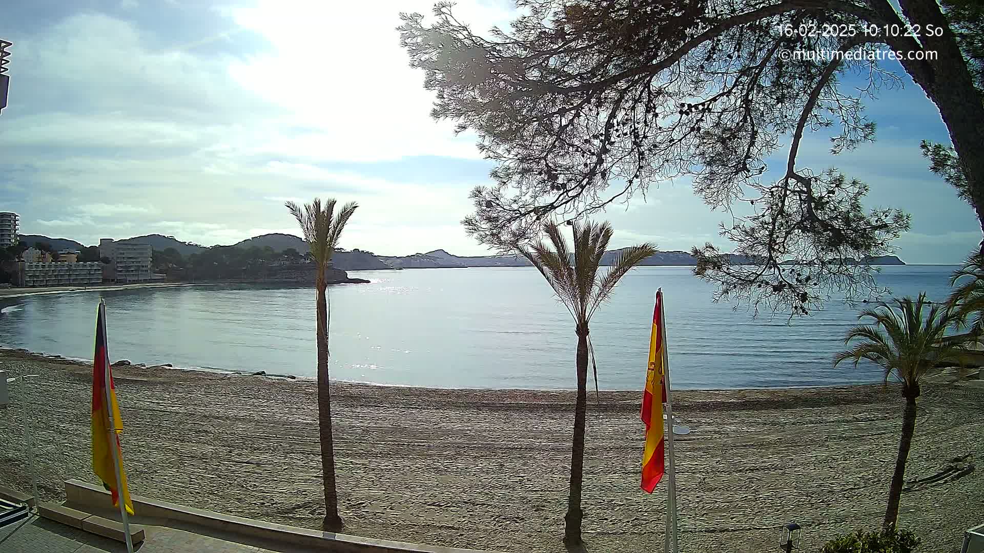 A partly sunny day shows a sandy beach, calm sea, palm trees, and buildings along the coast.