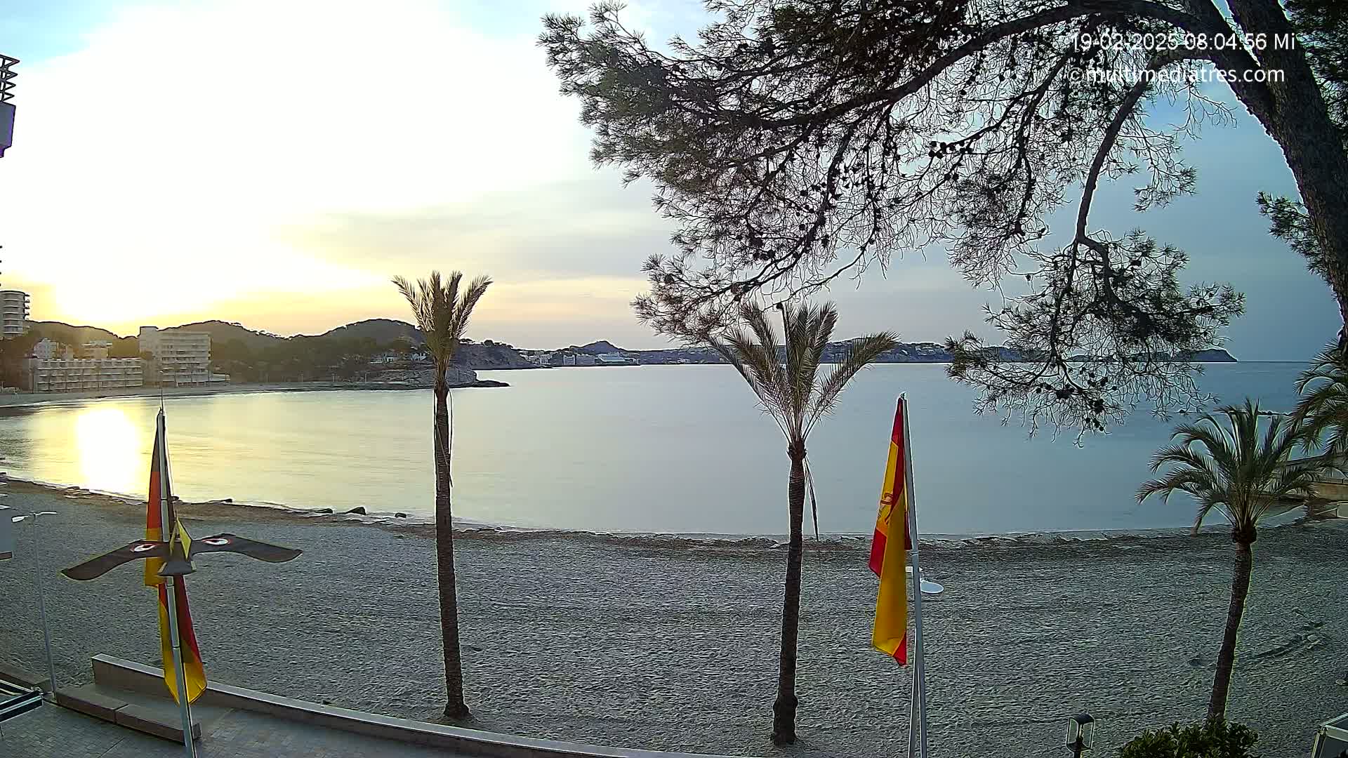 A calm bay at sunrise is viewed from a beach with palm trees and flags, under a partly cloudy sky.