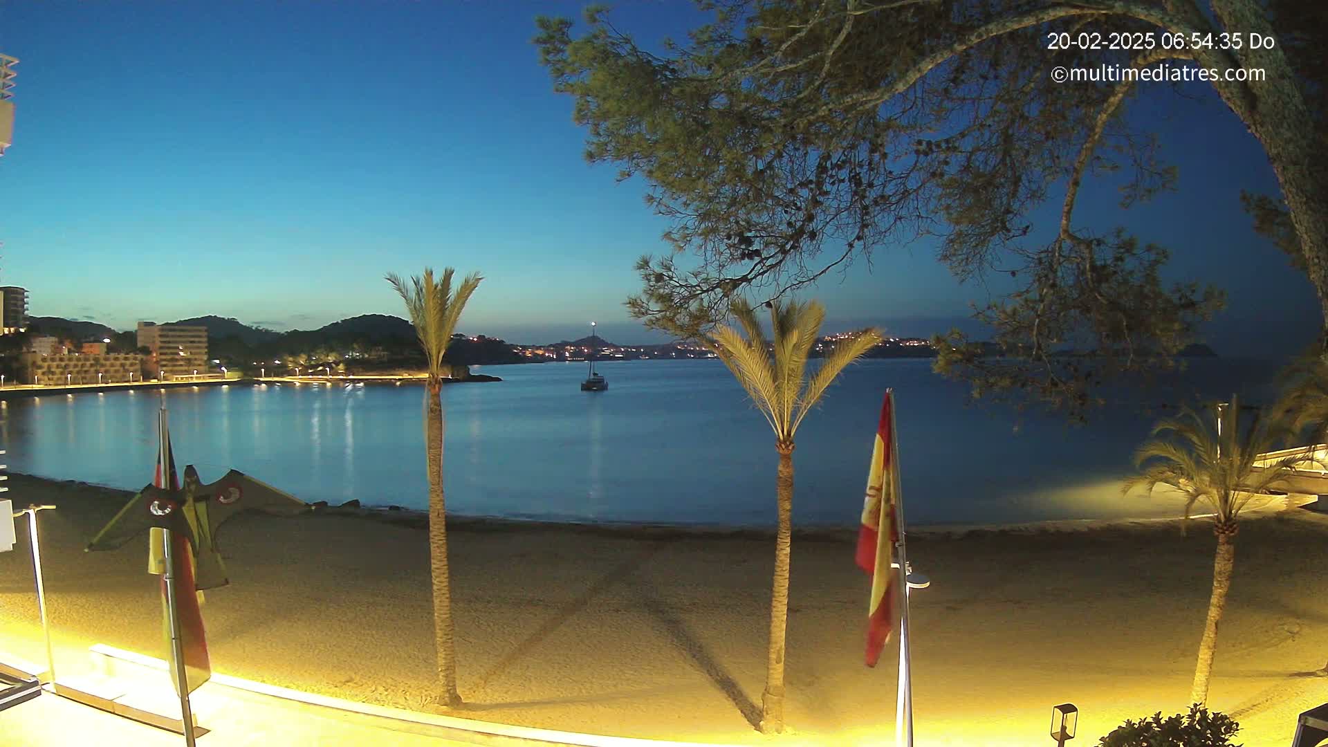 A sandy beach at twilight, bordered by palm trees and buildings, overlooks a calm bay with a single boat under a clear, dark blue sky.