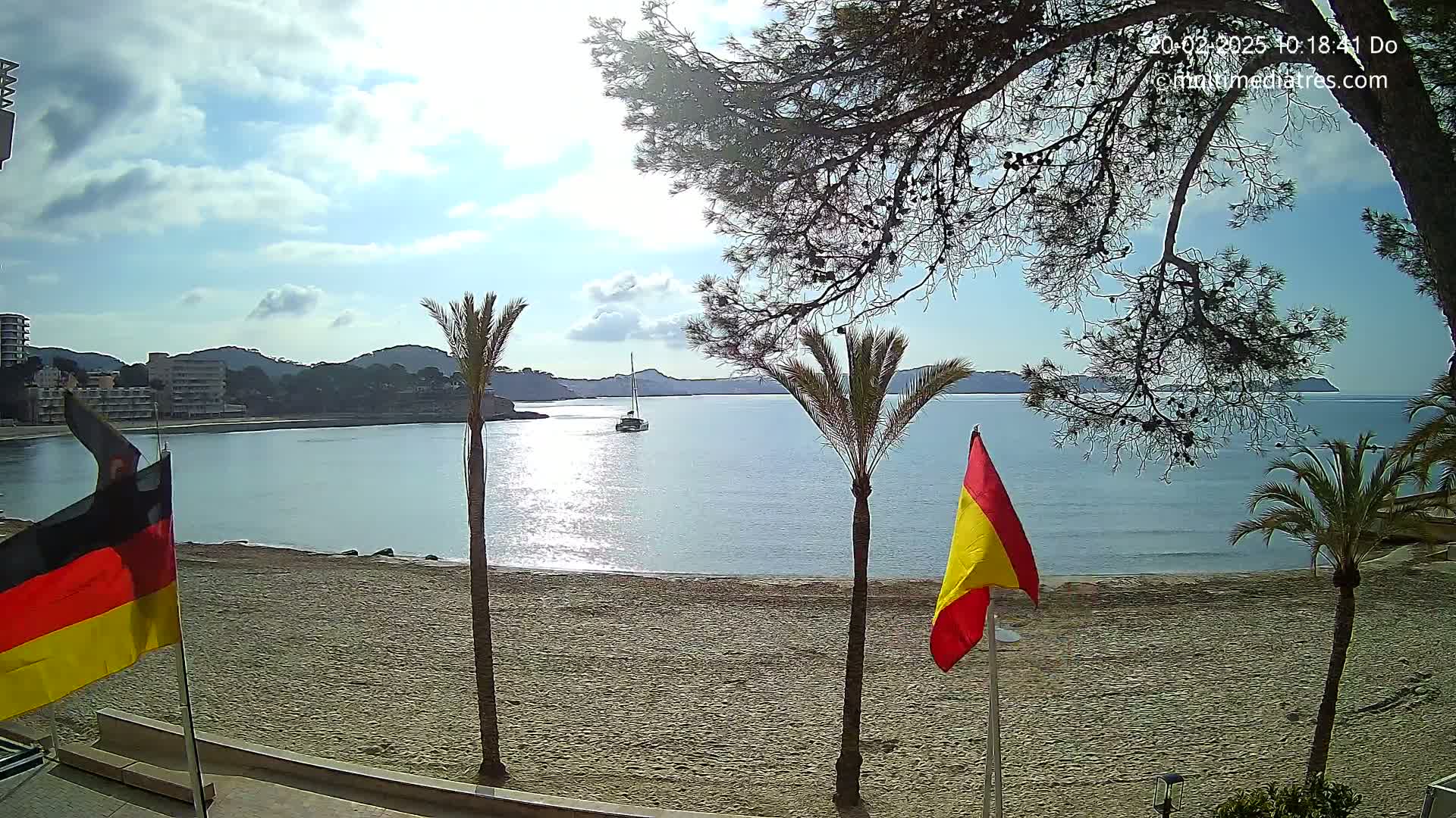 A sandy beach with palm trees and flags, overlooks a calm bay with a sailboat under a partly sunny sky.