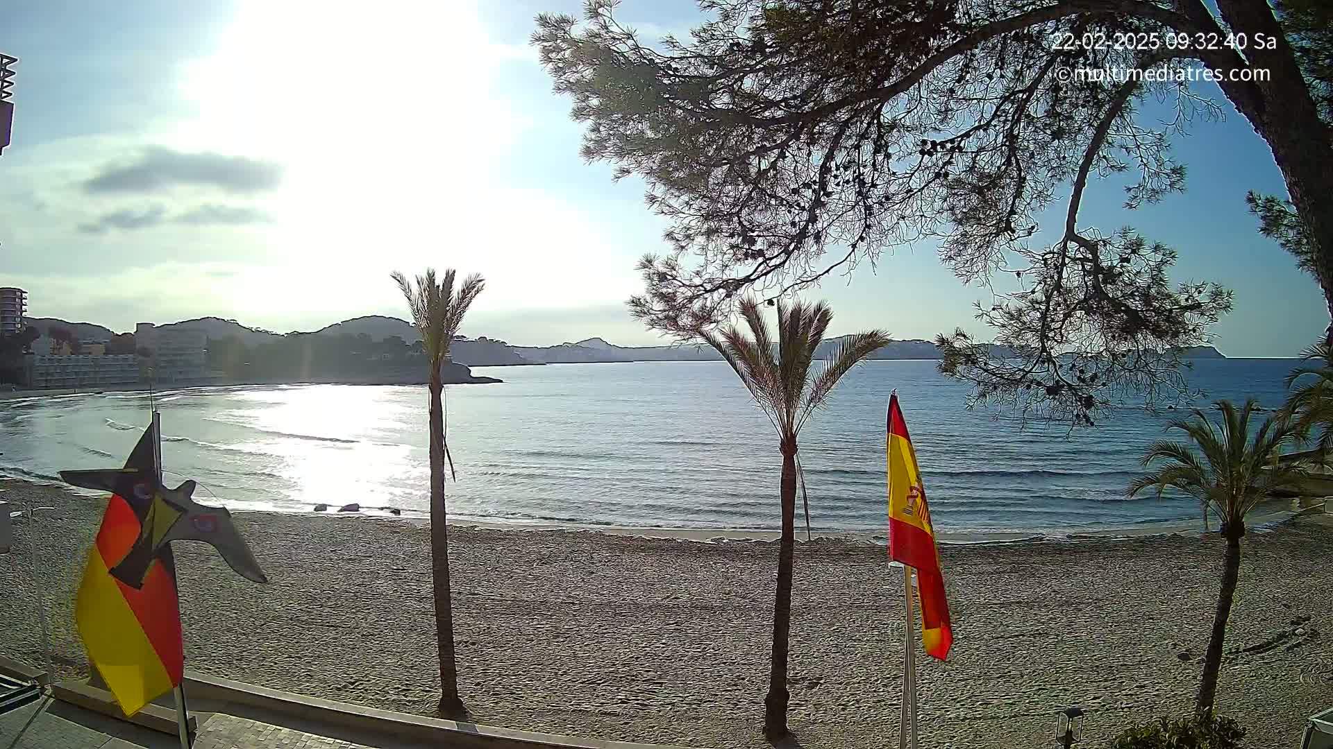 A sandy beach with calm, clear water, palm trees, and a partly sunny sky overlooks a bay with hills in the background.