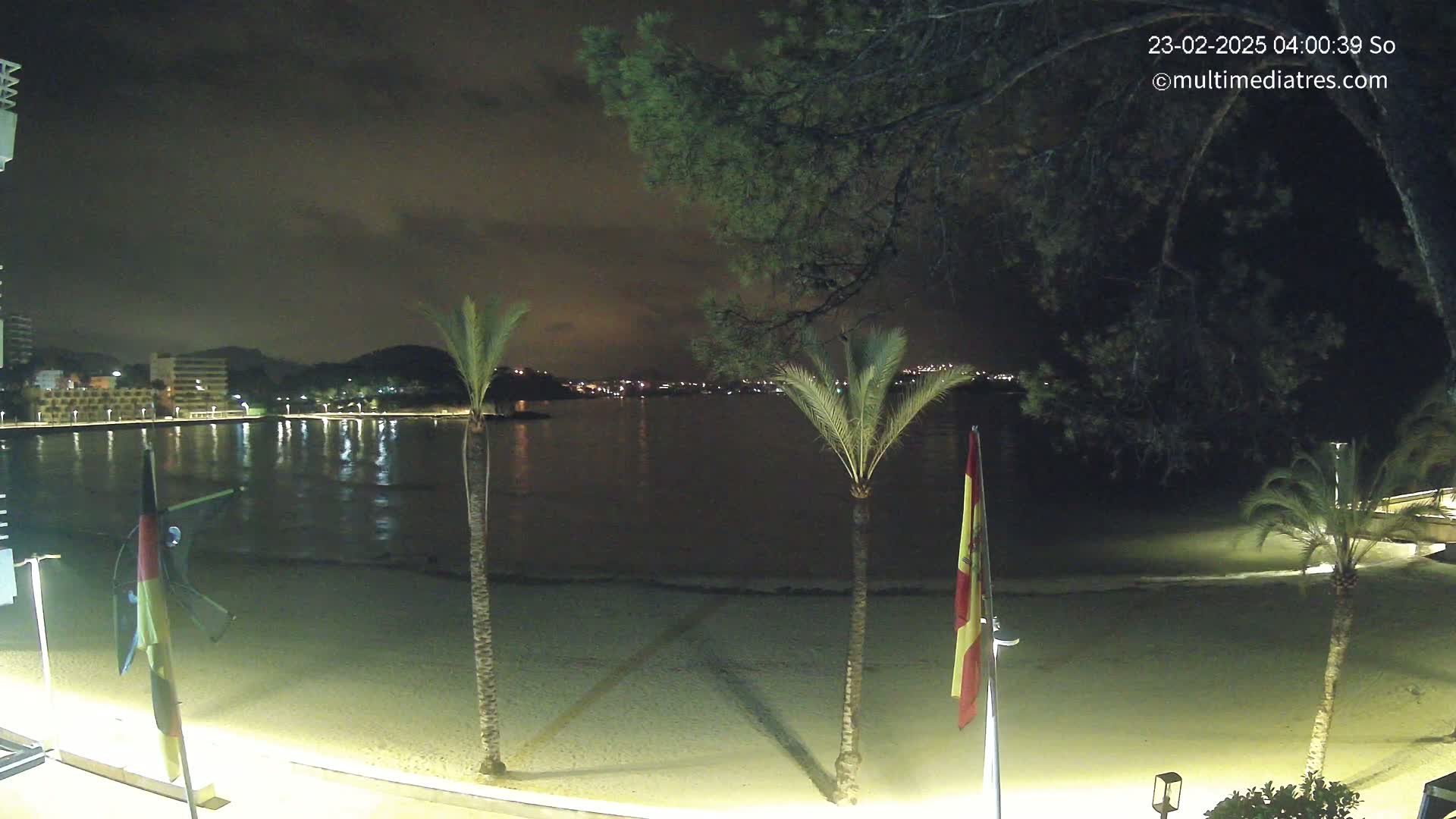 A nighttime view of a sandy beach, calm water, and a city skyline across the water, illuminated by lights, under a dark, cloudy sky.