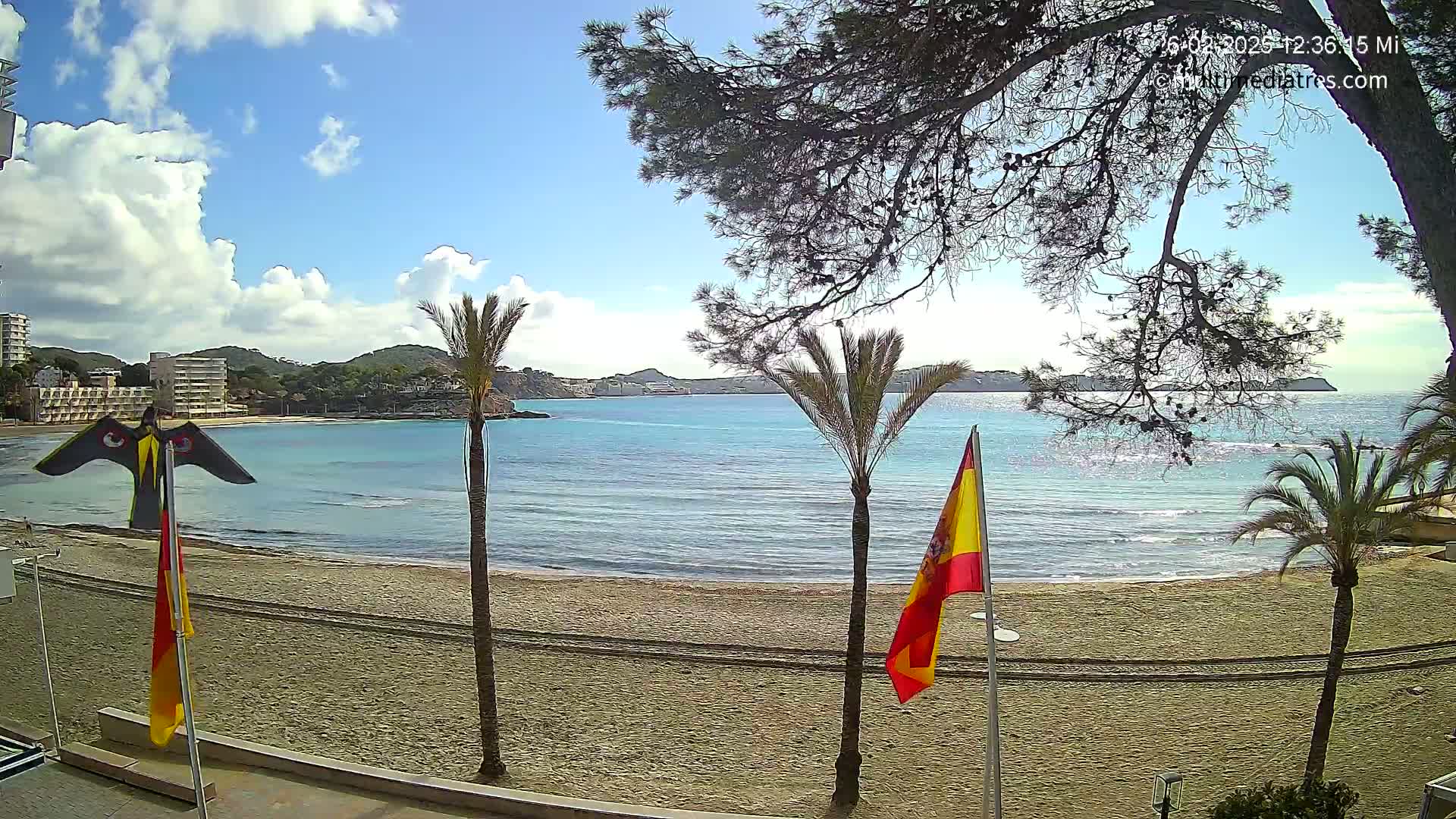 A sandy beach with calm, turquoise water, palm trees, and a Spanish flag is visible under a partly cloudy sky.