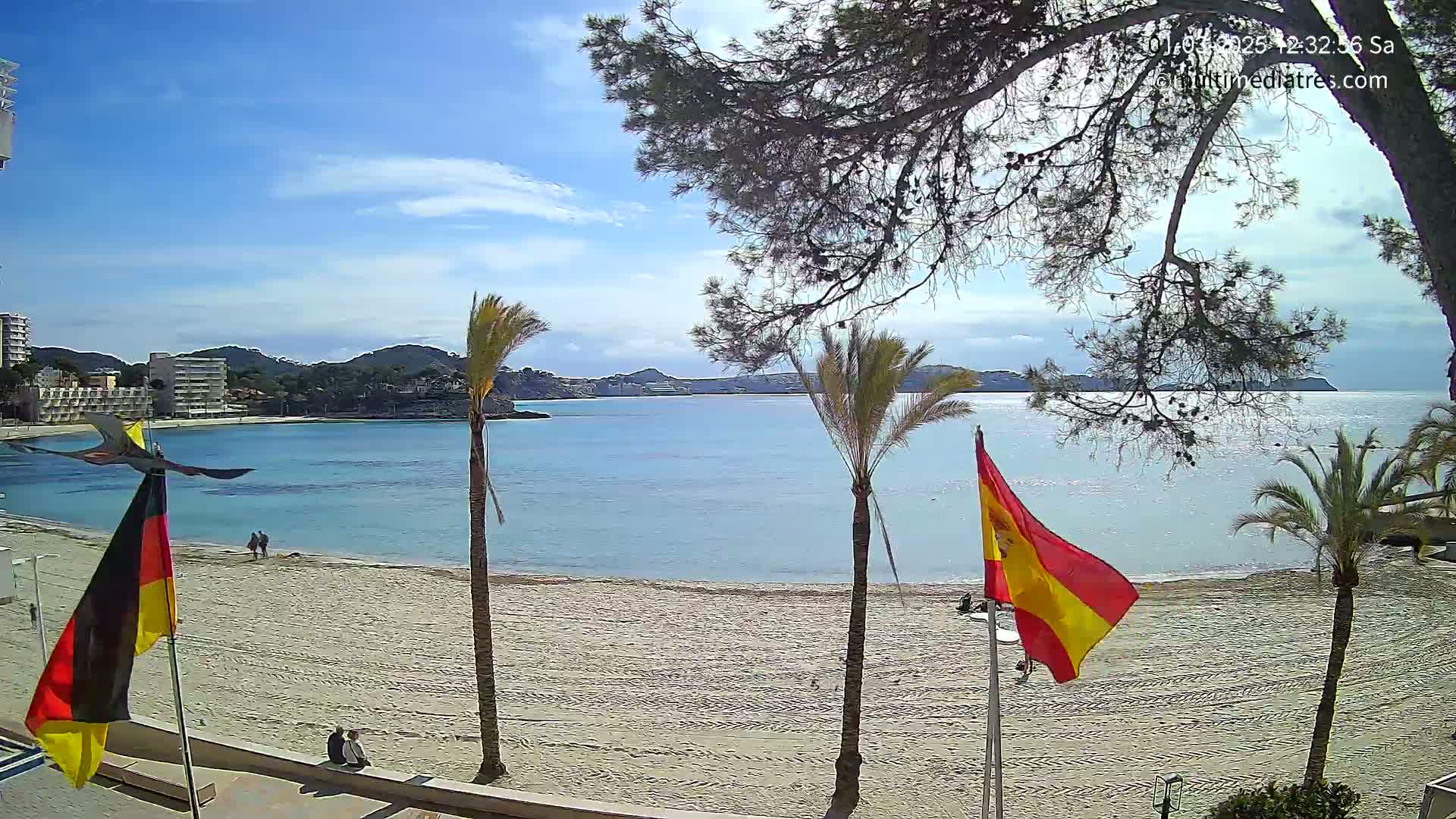 A sandy beach with calm, light-blue water and palm trees under a partly sunny sky is shown, with flags of Germany and Spain on the left and right.