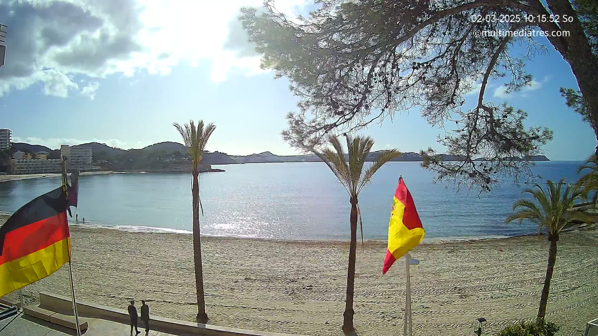 A sandy beach with palm trees and two flags, one German and one Spanish, overlooks a calm bay under a partly sunny sky.
