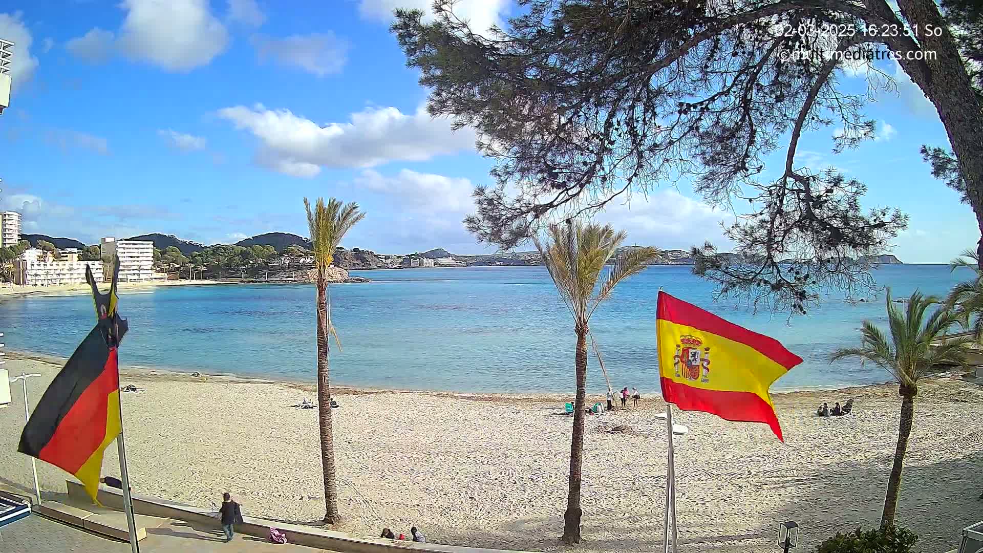 A sandy beach with calm, clear blue water, palm trees, and flags of Germany and Spain under a partly sunny sky.