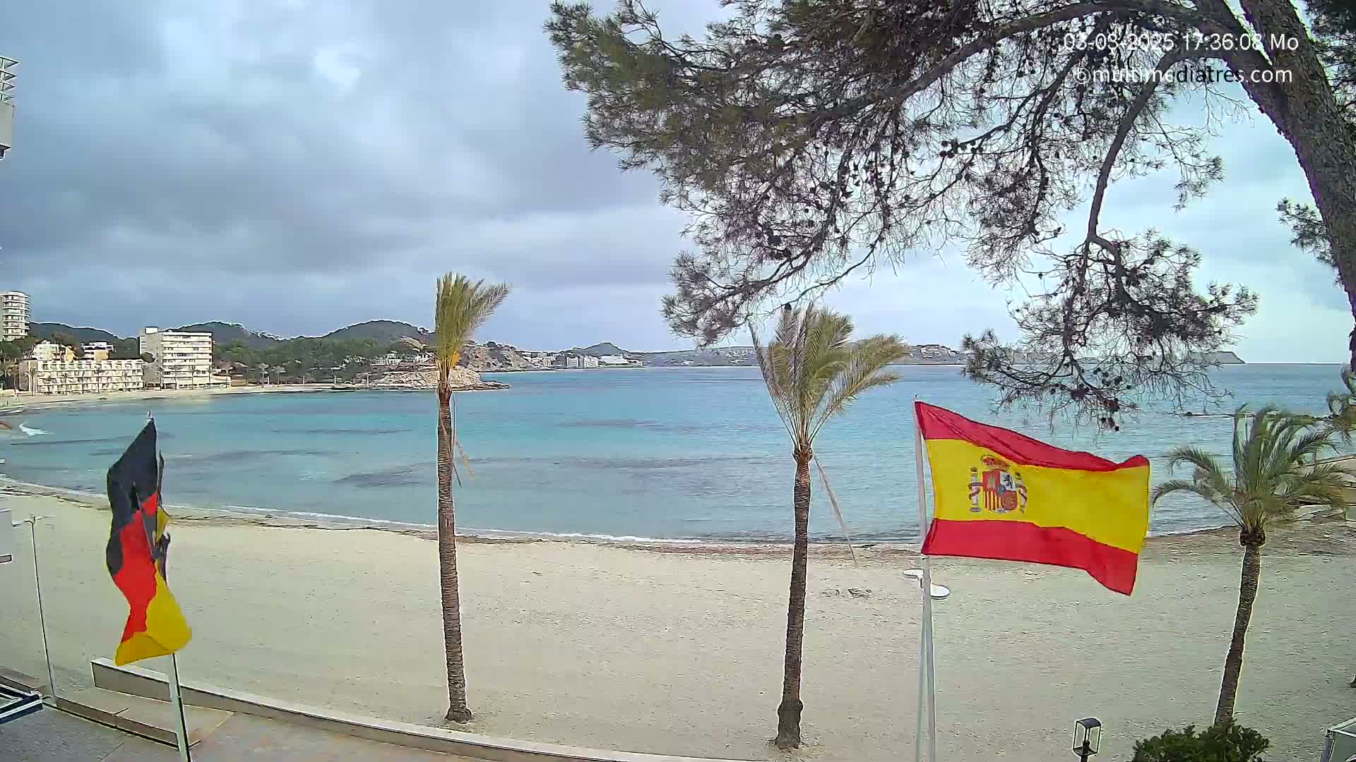 A sandy beach with calm, turquoise water is viewed from a beachfront vantage point under a cloudy sky, with Spanish and German flags waving in the breeze, and palm trees and buildings visible in the background.