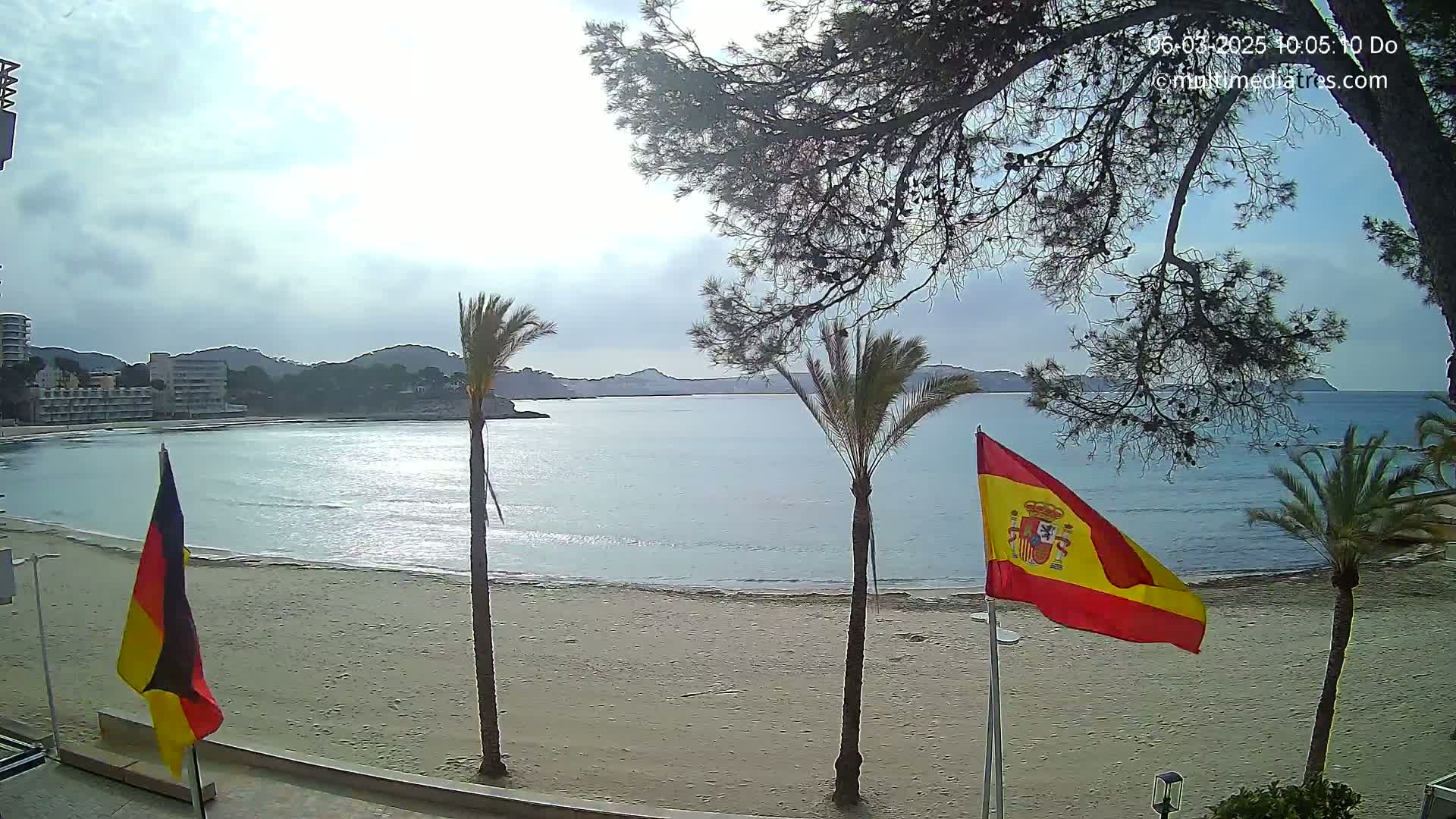 A sandy beach with calm water, palm trees, and flags of Spain and Germany is visible under an overcast sky.