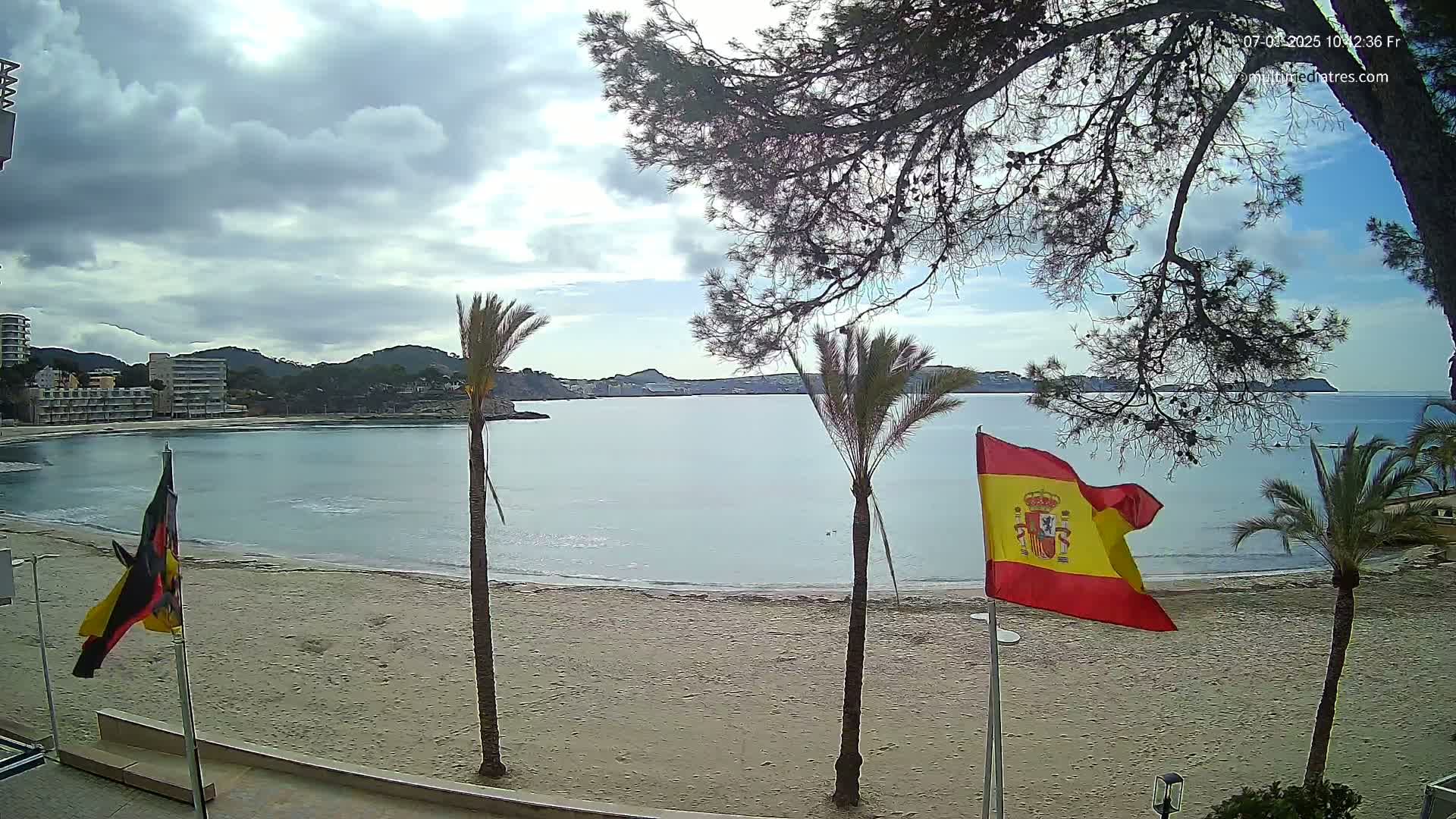 A sandy beach with calm water and buildings in the background under a partly cloudy sky, with palm trees and flags in the foreground.