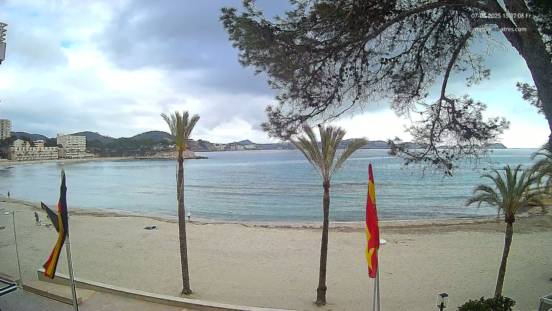 A sandy beach with calm, blue water stretches out to hills under a cloudy sky, palm trees and flags standing along the shoreline.