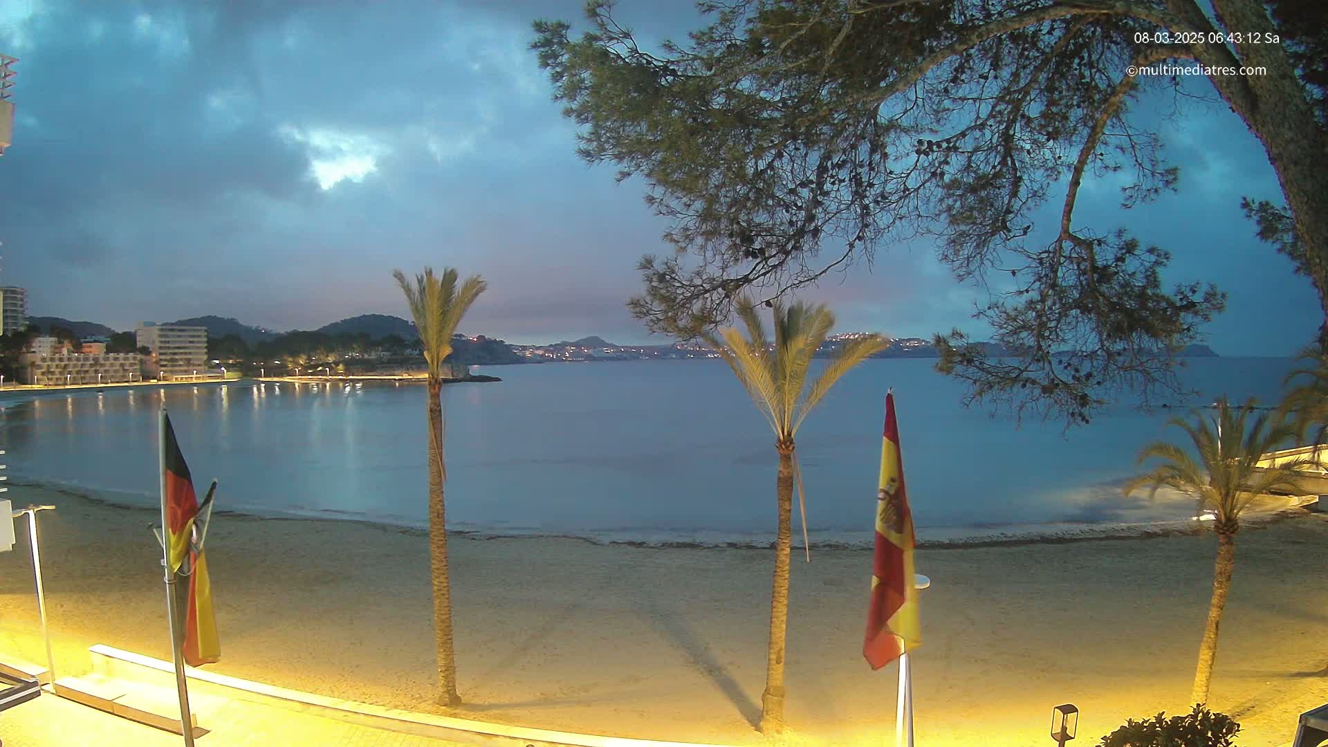 A sandy beach at twilight is illuminated by lights, with calm water, palm trees, and flags visible, under a partly cloudy sky.