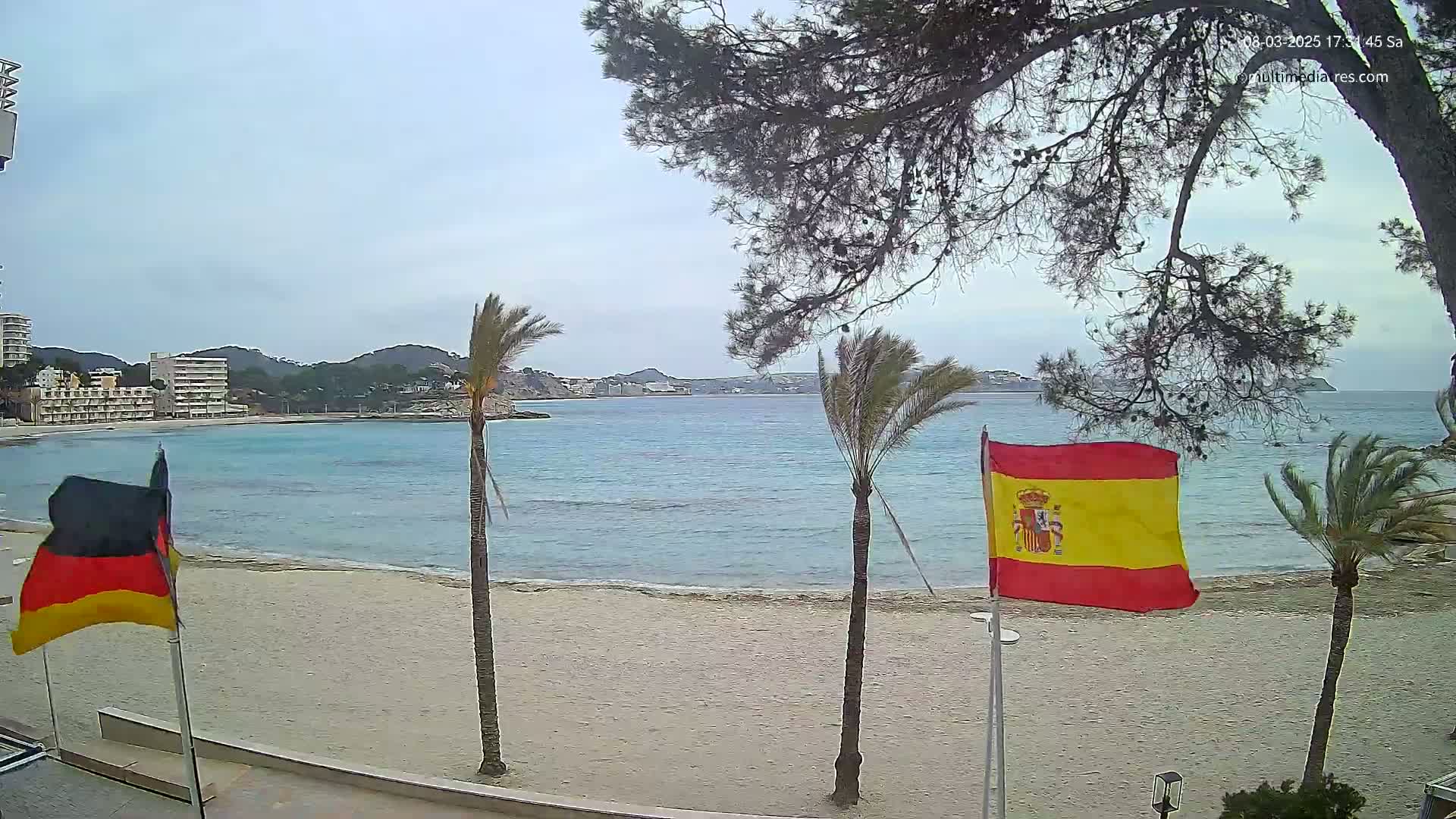 A sandy beach with calm, turquoise water stretches out before buildings and hills, under an overcast sky, with German and Spanish flags waving in the breeze near palm trees.