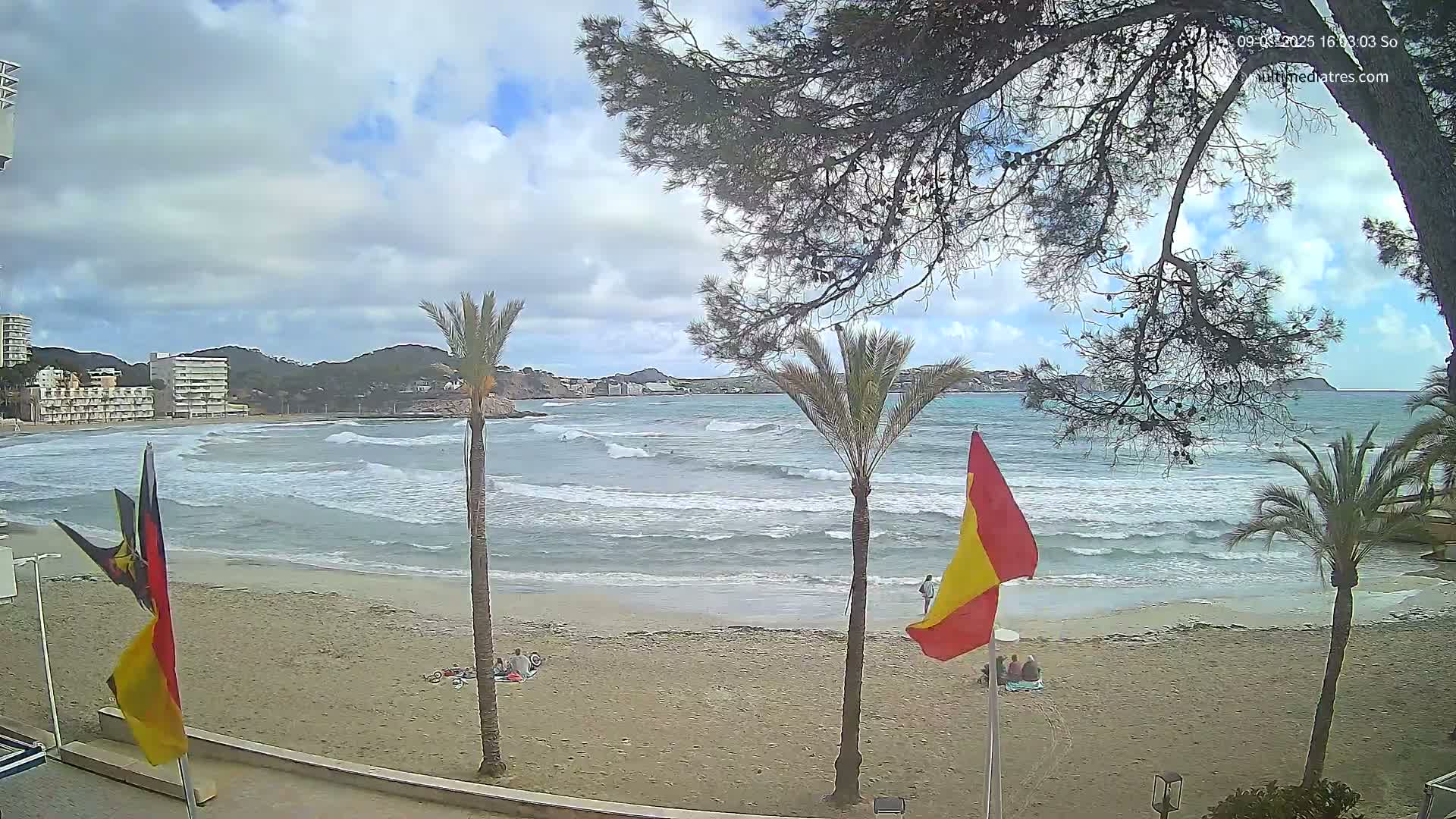 A sandy beach with moderate waves, palm trees, and a few people are visible under a partly cloudy sky.