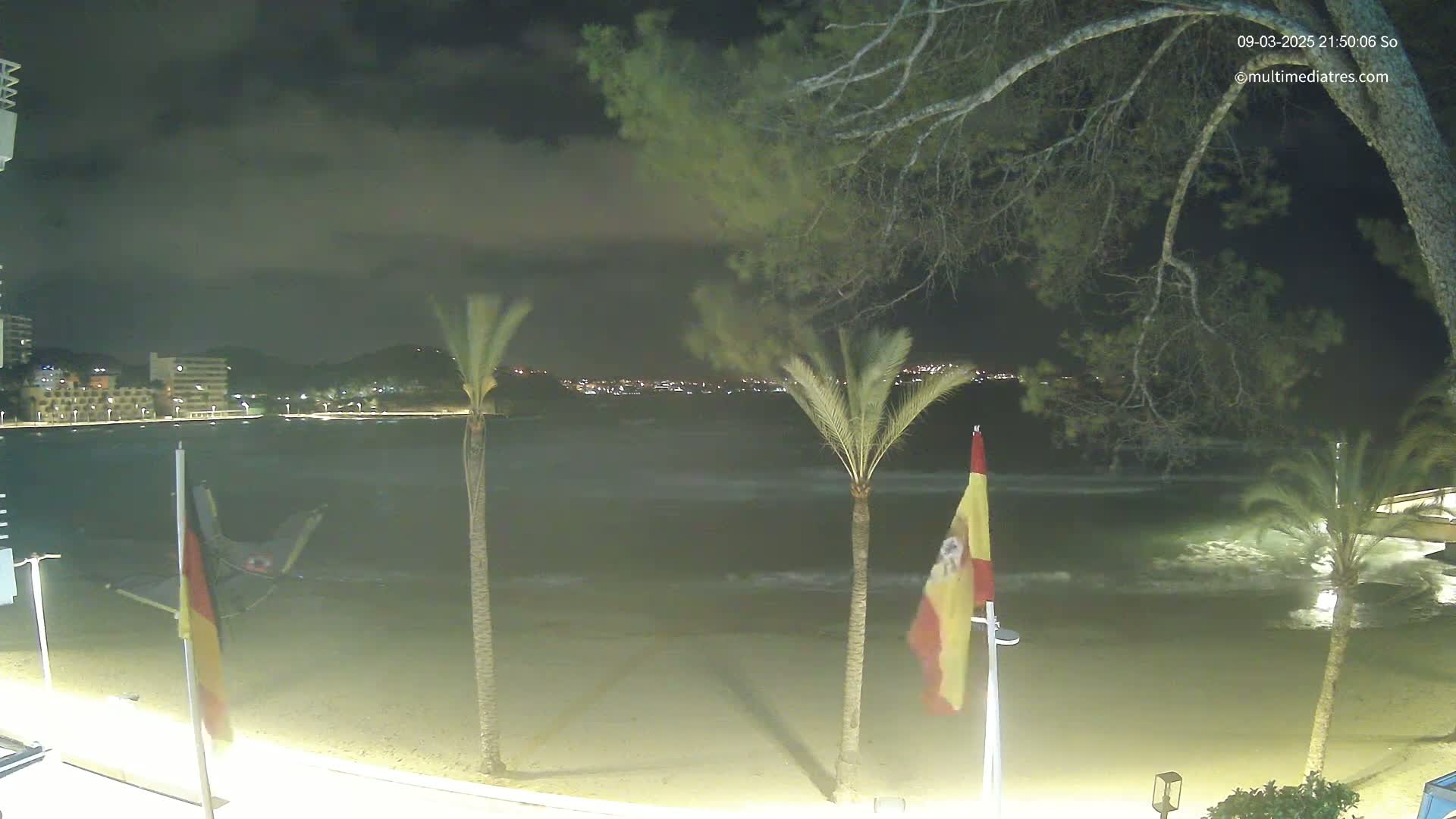 A nighttime beach scene shows a calm sea with small waves lapping the shore under a partly cloudy sky, palm trees and flags in the foreground, and a city skyline in the distance.