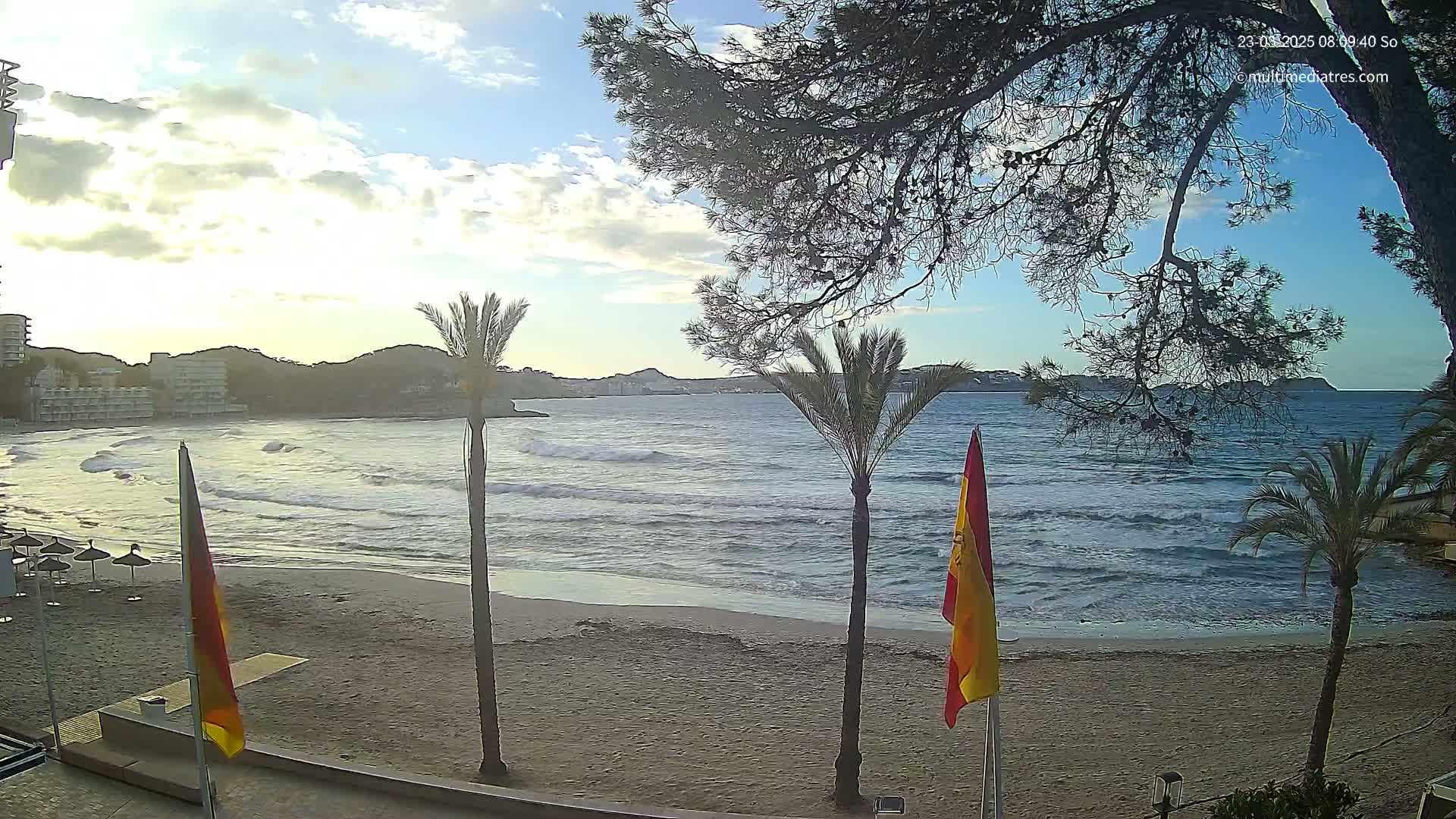 A sandy beach with palm trees and gently rolling waves under a partly cloudy sky is visible, with buildings and hills in the distance.