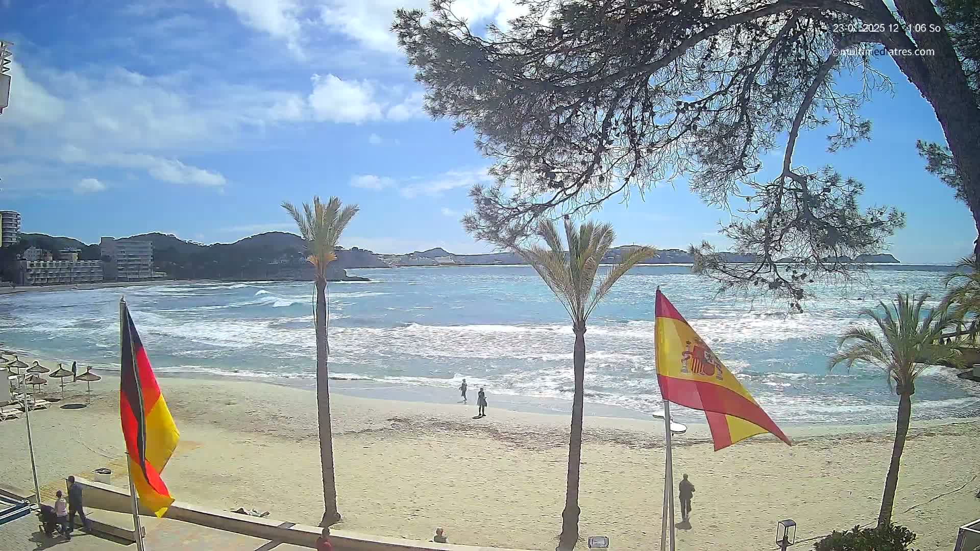A sandy beach with some people on it, bordered by palm trees and buildings, overlooks a wavy ocean under a partly sunny sky.