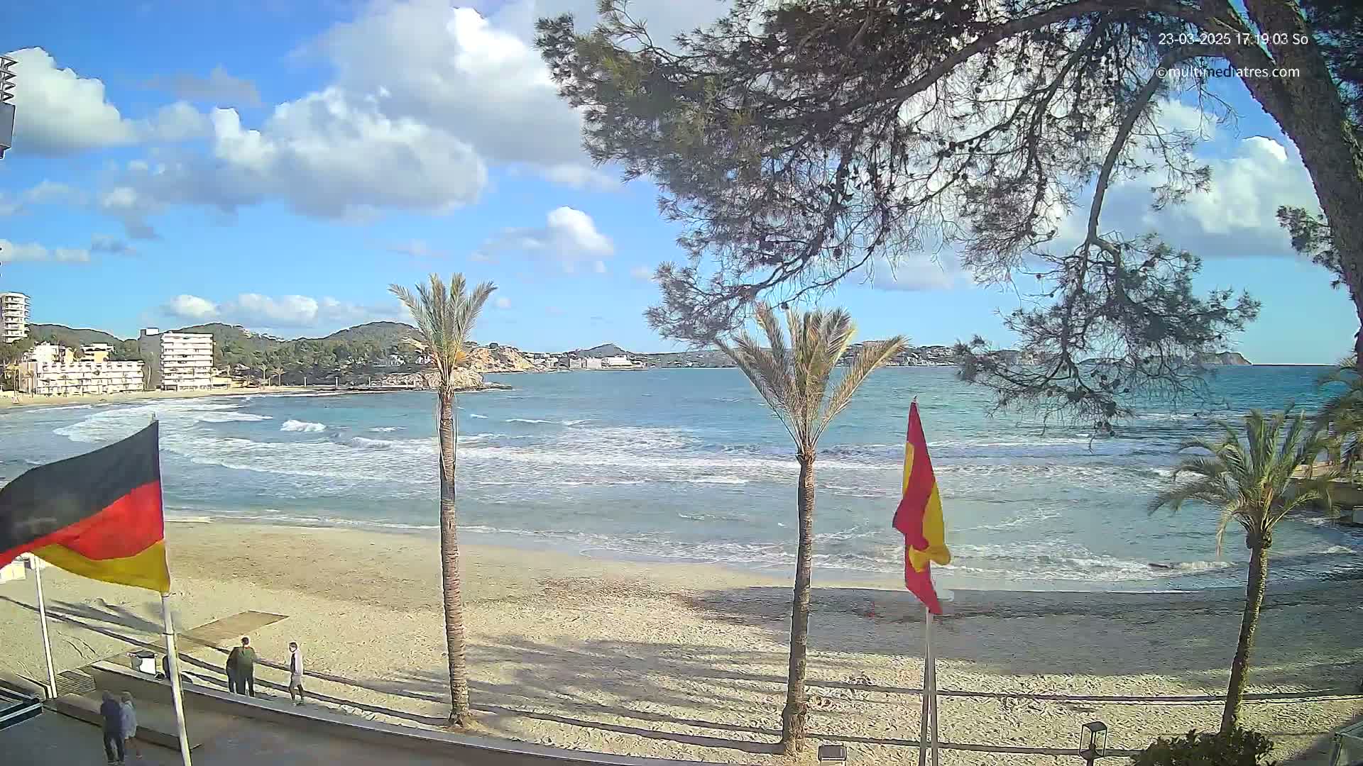 A partly cloudy sunny day at a sandy beach with calm waves, palm trees, and buildings in the background.