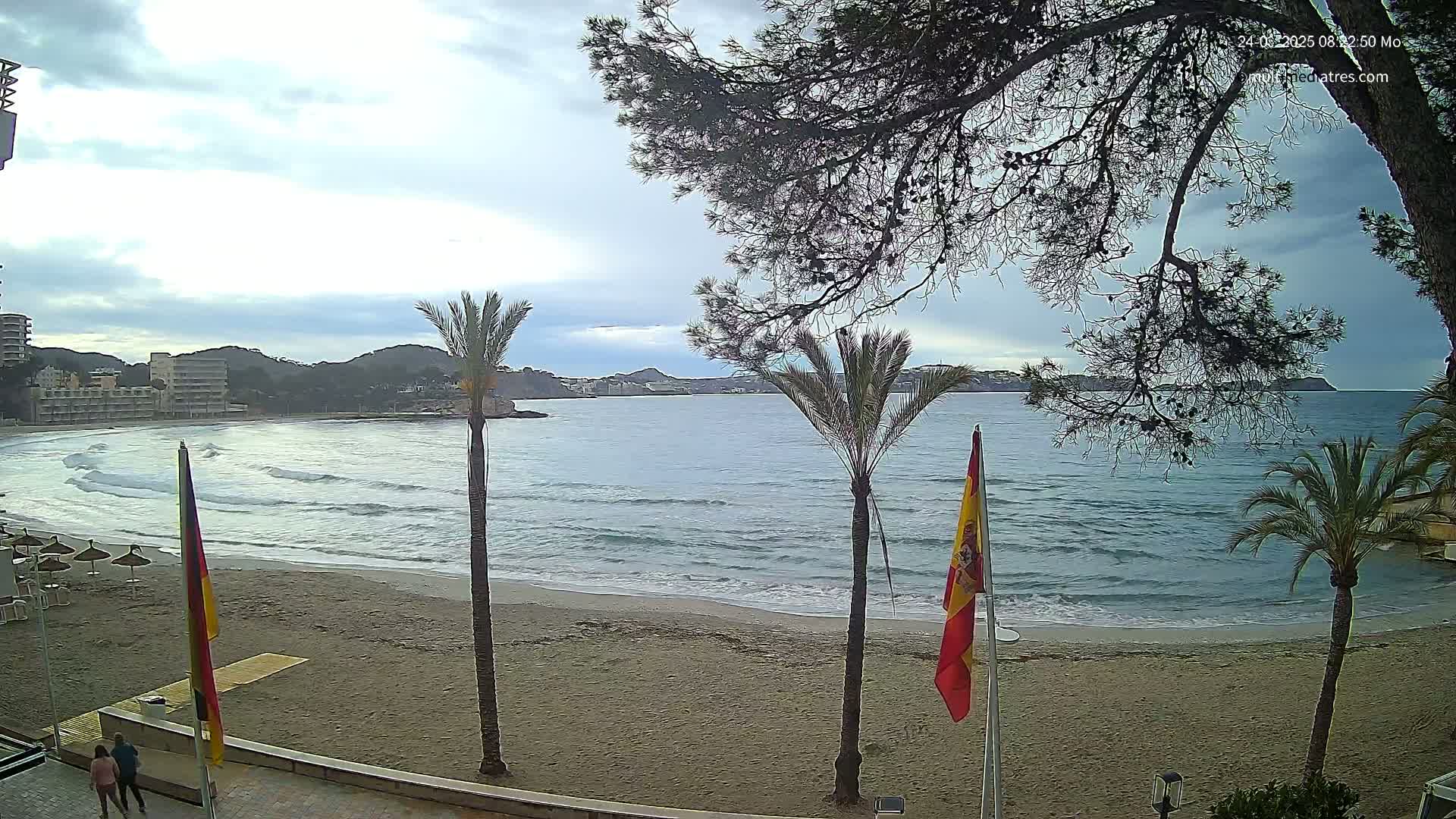 A sandy beach with gentle waves laps the shore under a cloudy sky, palm trees and buildings are visible in the background.