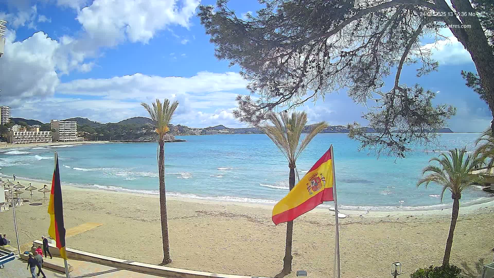A sandy beach with calm, turquoise water stretches out under a partly cloudy blue sky, flanked by palm trees and buildings, with Spanish and German flags displayed nearby.