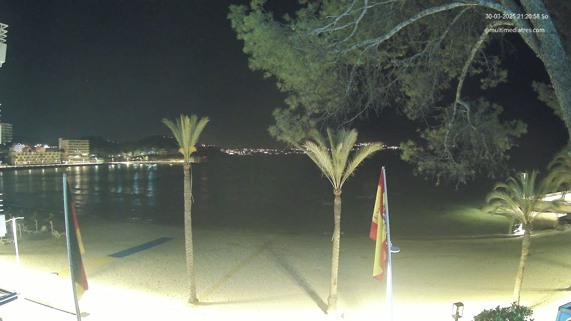 A nighttime view of a sandy beach with palm trees and flags, overlooking a calm body of water and a distant, illuminated town.