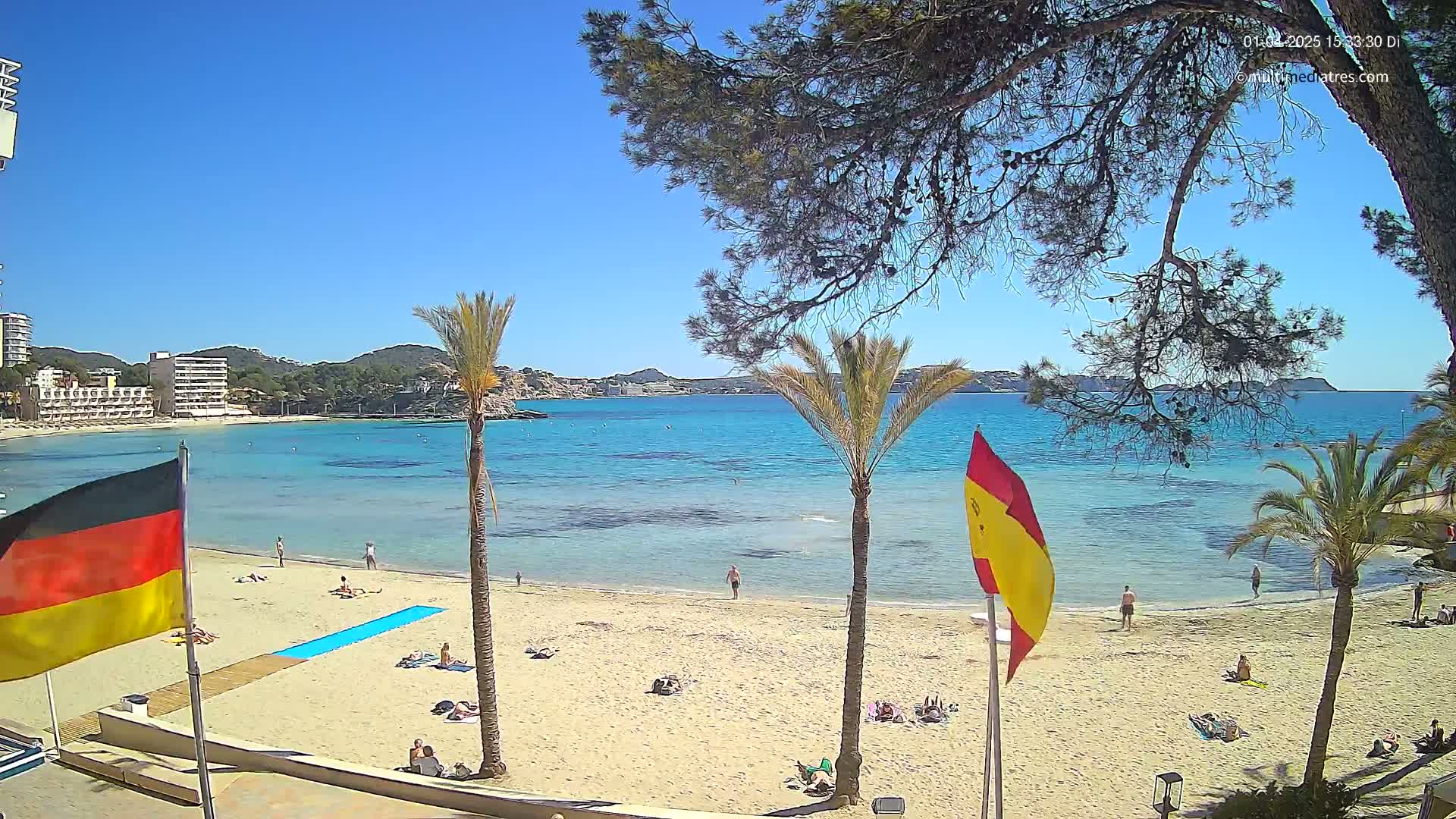 A sandy beach with people sunbathing and swimming in calm, clear turquoise water under a bright sunny sky, framed by palm trees and buildings in the distance.