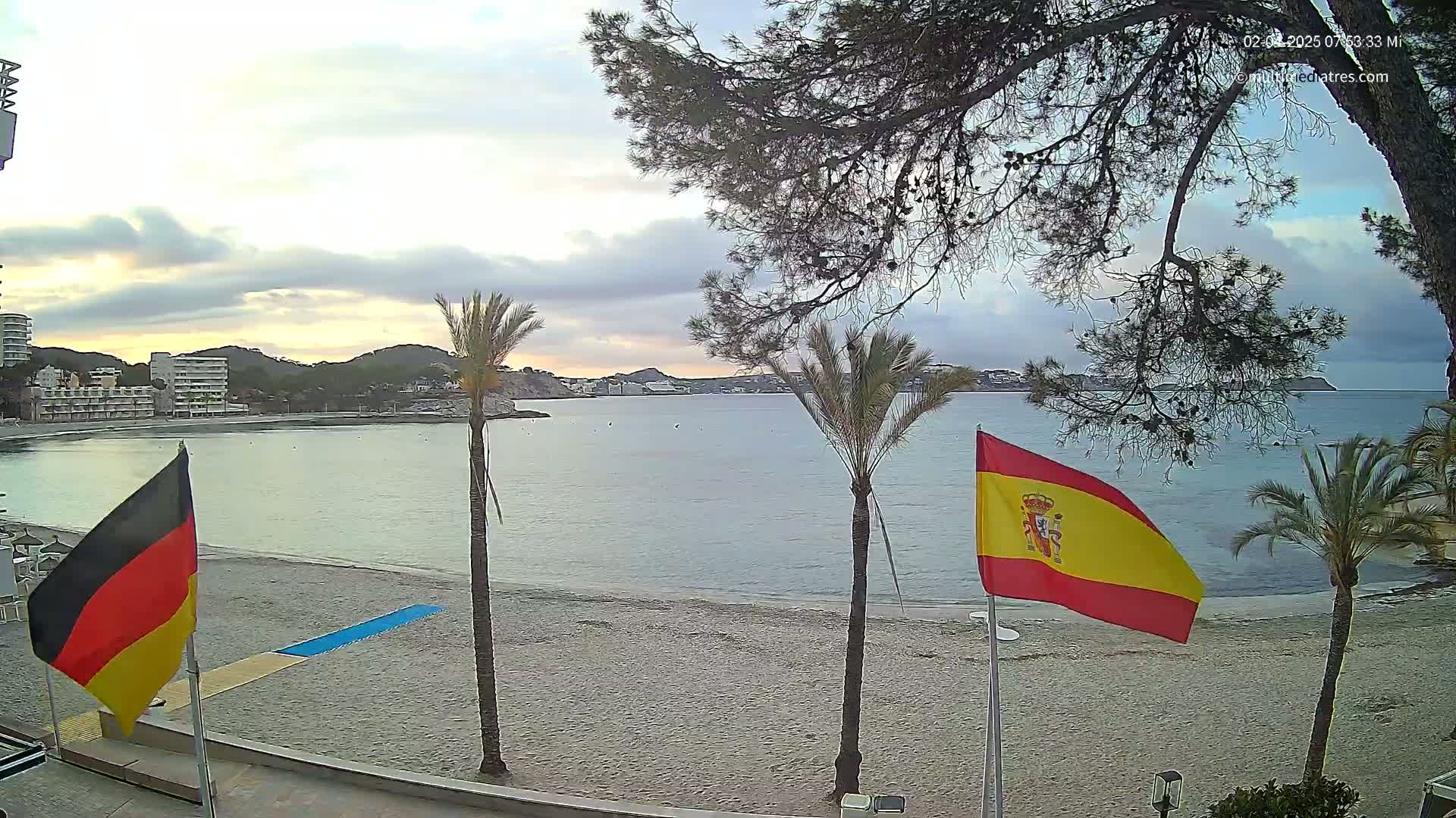 A sandy beach with calm water, palm trees, and flags of Germany and Spain is visible under a partly cloudy sky.