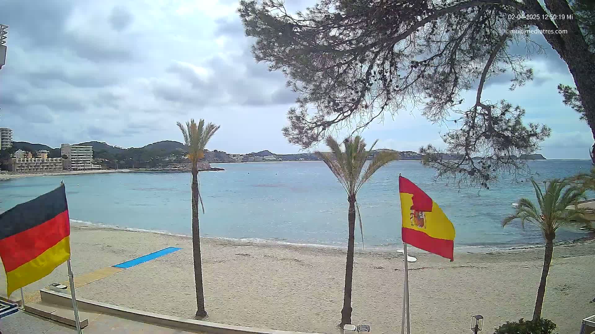 A sandy beach with palm trees and flags, overlooks a calm, light blue bay under a partly cloudy sky.