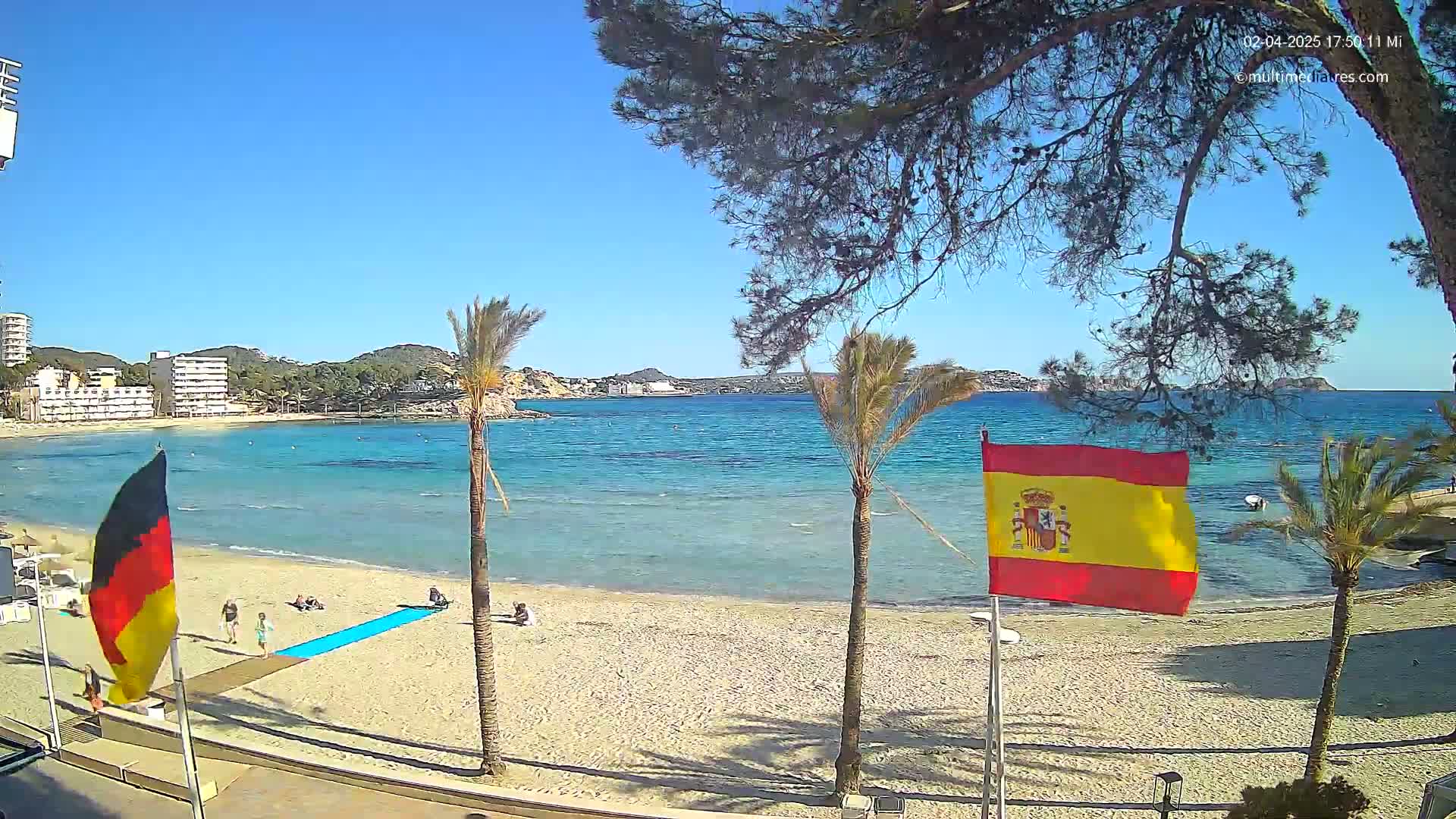 A sandy beach with calm, clear turquoise water is bordered by palm trees, buildings, and flags of Germany and Spain under a sunny sky.