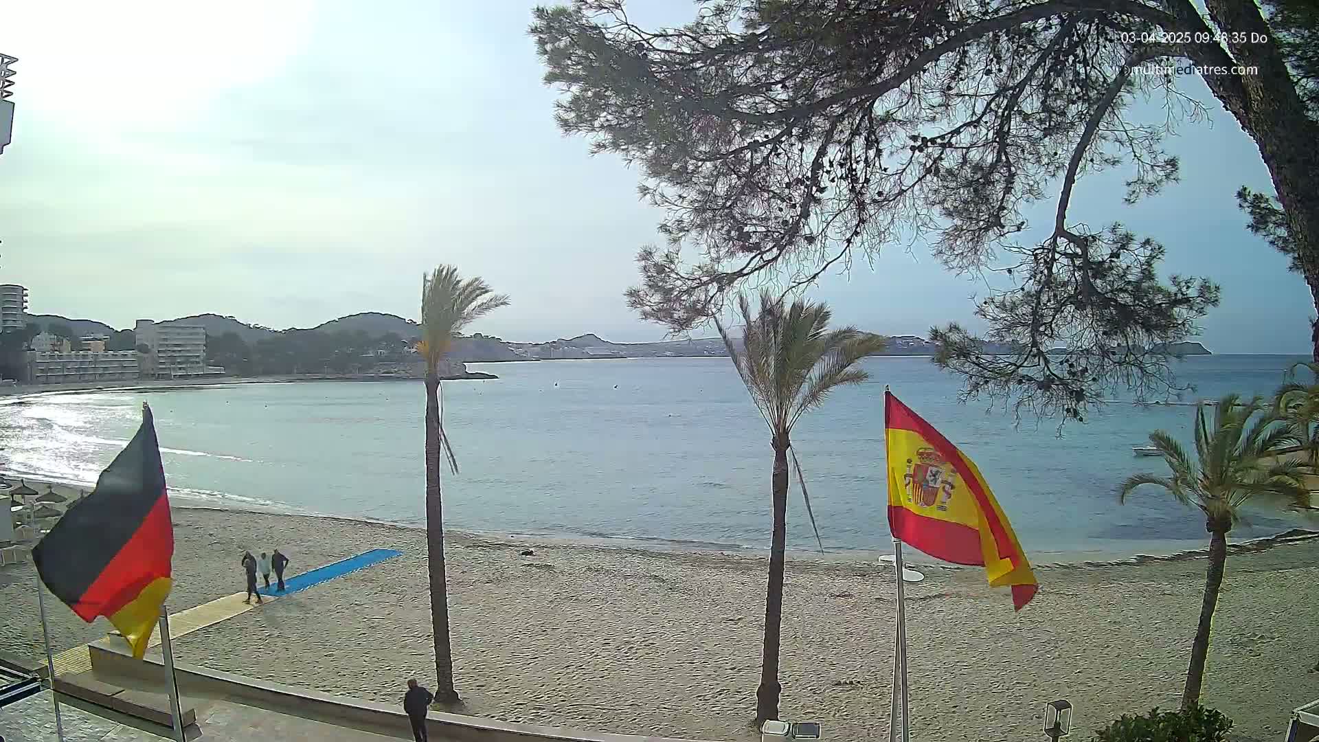 A sandy beach with calm, blue water and palm trees under a mostly sunny sky, featuring German and Spanish flags.