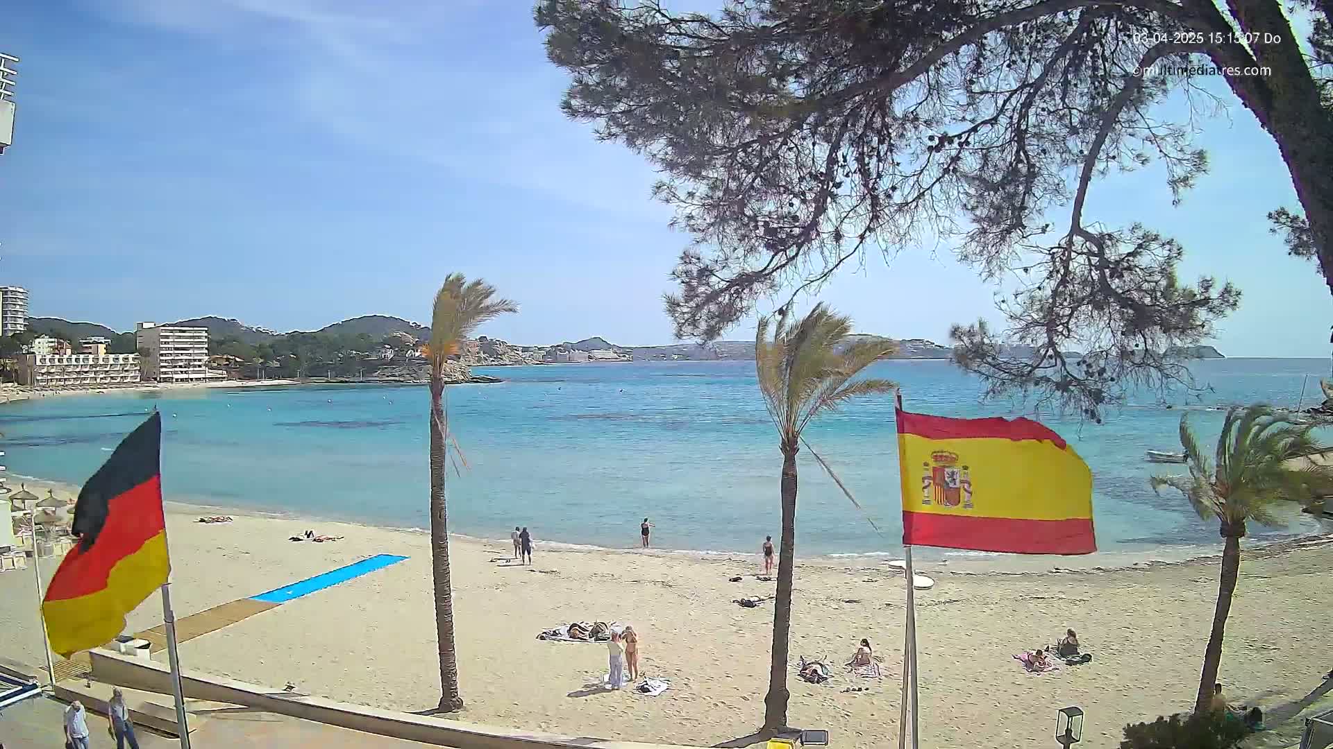 A sandy beach with several people scattered across it, palm trees, and flags of Spain and Germany, overlooks a calm turquoise bay under a bright sunny sky.