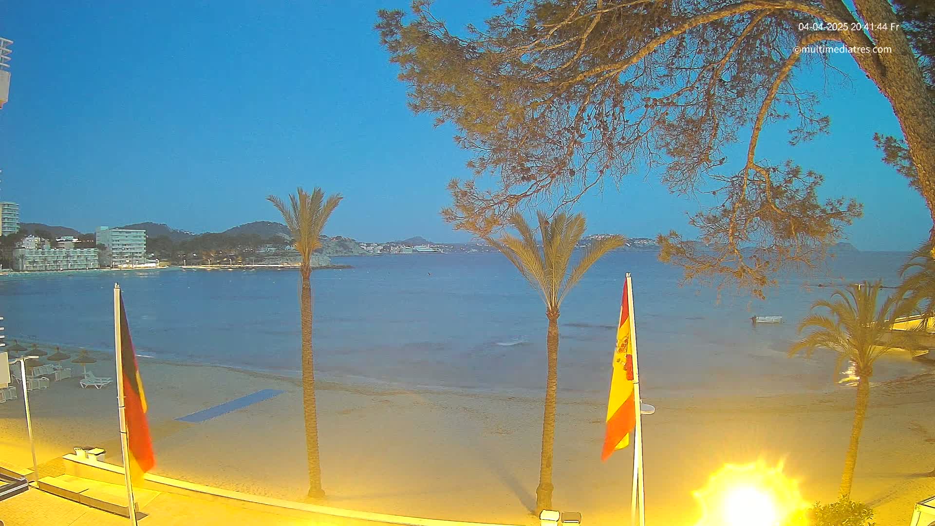 A sandy beach at dusk, under a clear blue sky, is bordered by calm water, palm trees, and buildings in the distance.