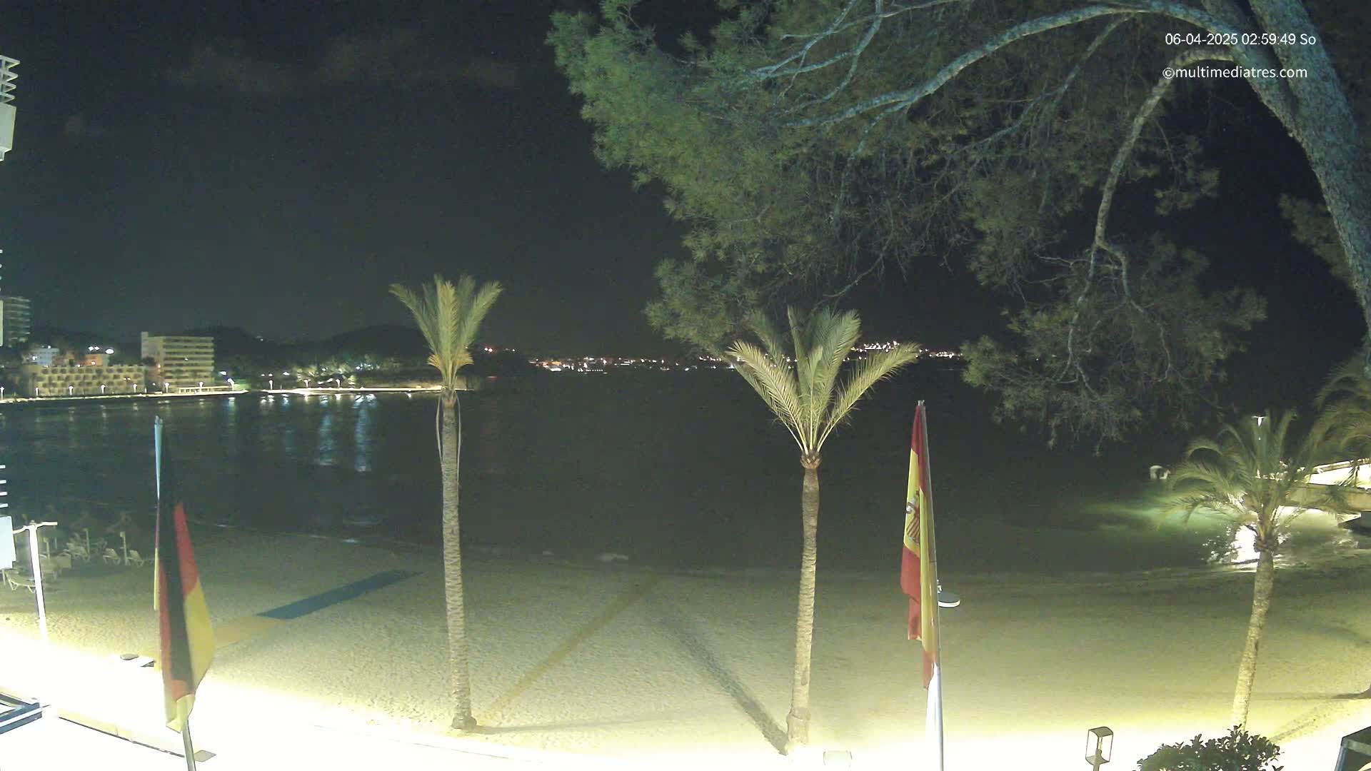 A nighttime view of a sandy beach with calm water, palm trees, and flags, overlooking a town illuminated with lights across the bay.